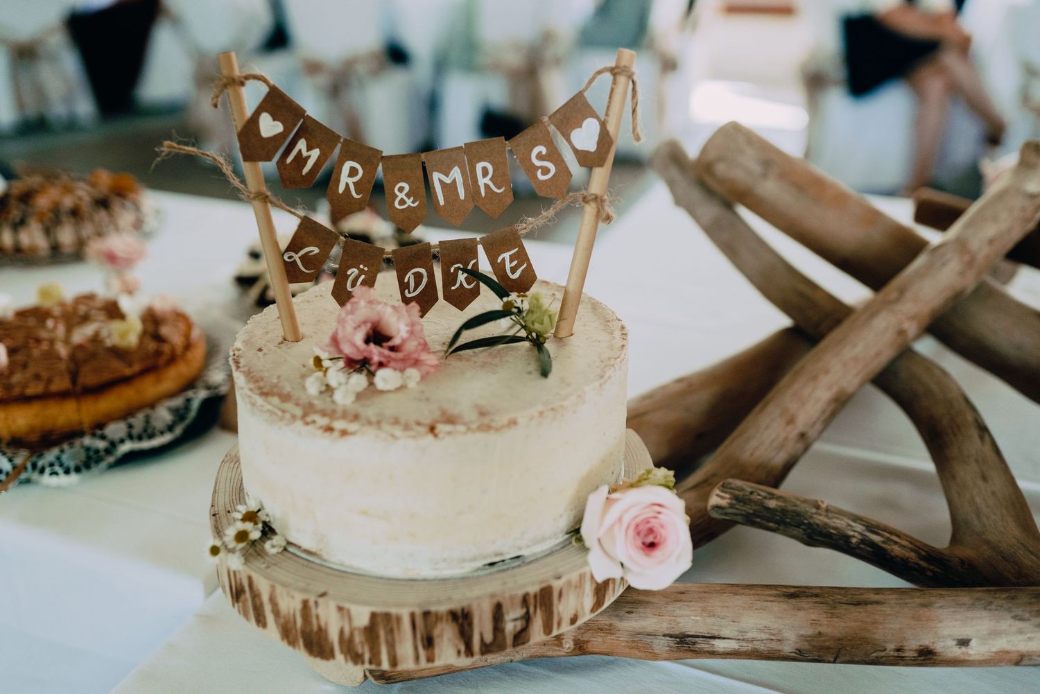 Rustic wedding cake on wooden slice with 'forever' bunting topper and pink rose decoration.