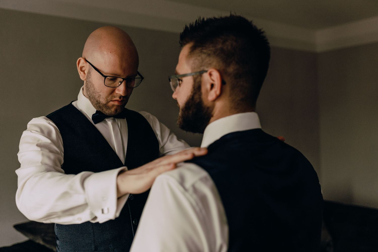 Two formally dressed men in white shirts and vests help each other prepare, one adjusting the other's attire.