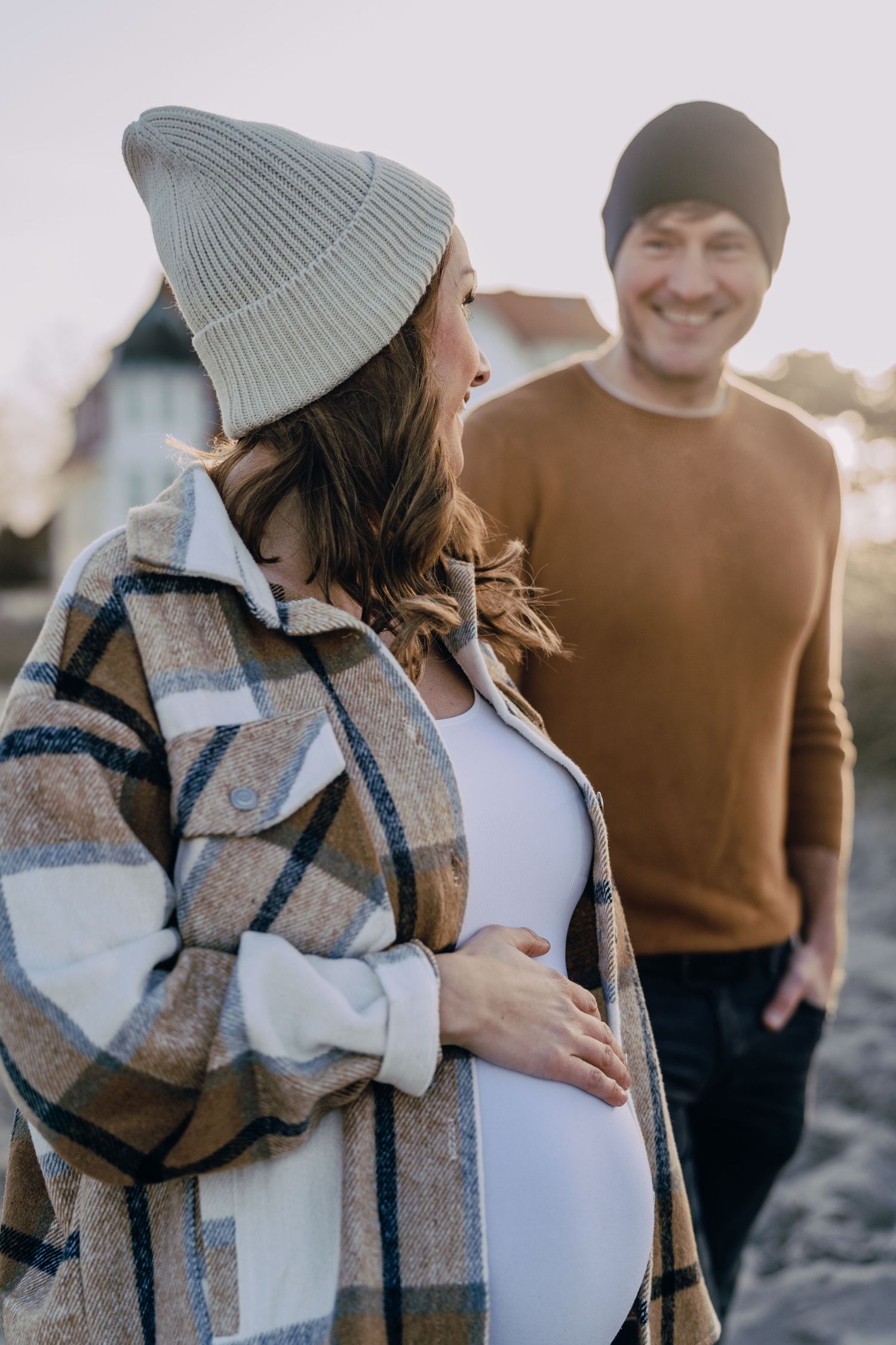A couple in cozy winter attire including a plaid shacket and knit beanie embraces during a tender outdoor photoshoot at sunset.