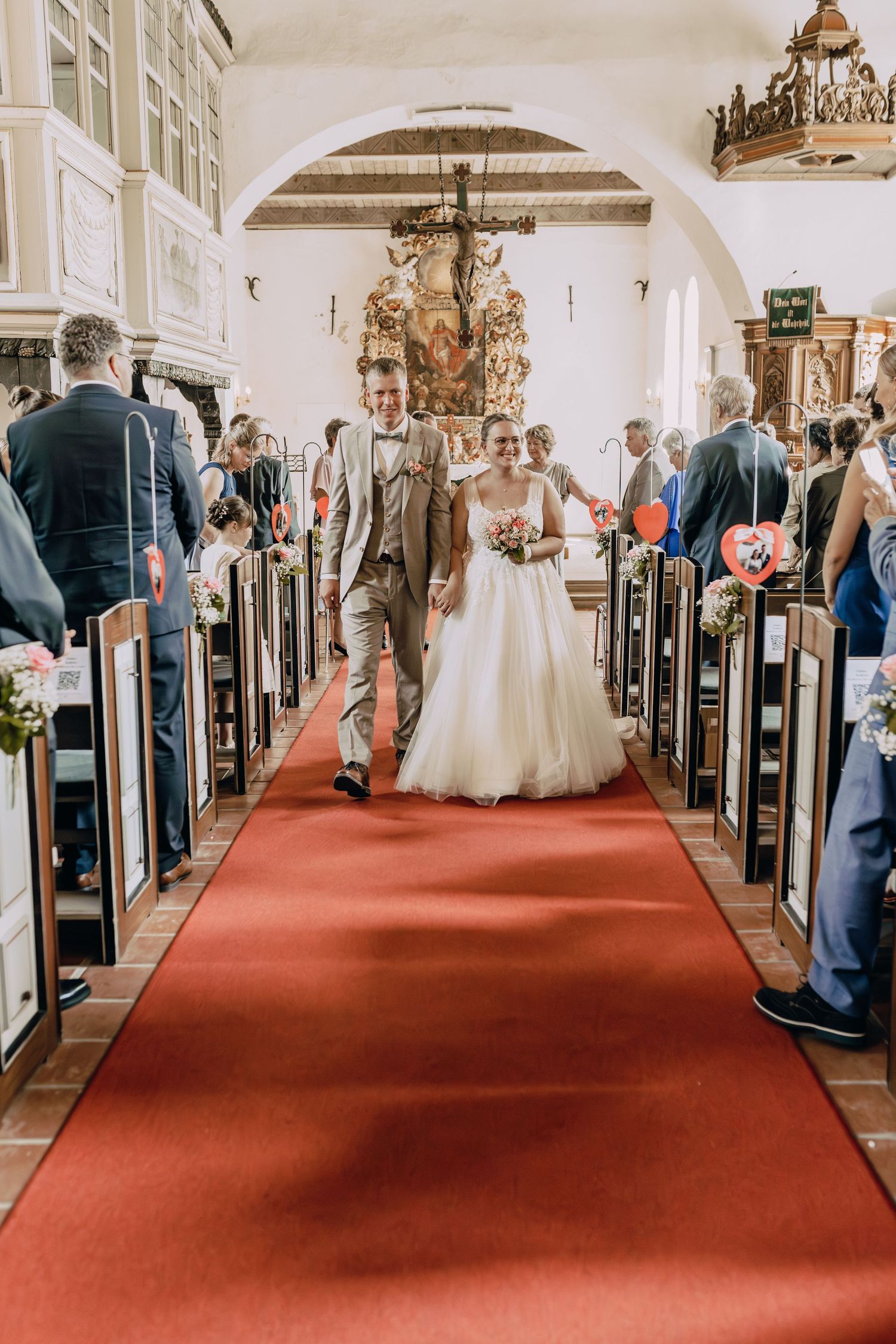 Wedding guests stand in church pews as bride and groom proceed down red carpeted aisle in traditional ceremony.