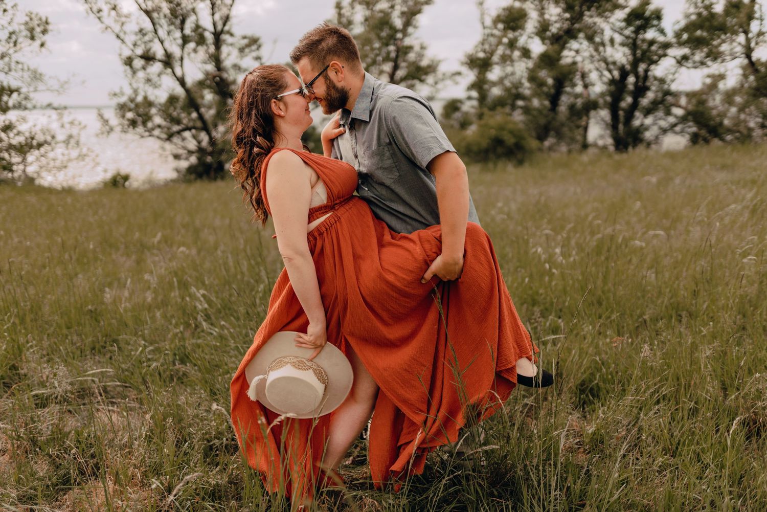 A couple in a romantic embrace stands in a grassy field at sunset, with the woman wearing a flowing red dress holding a hat.