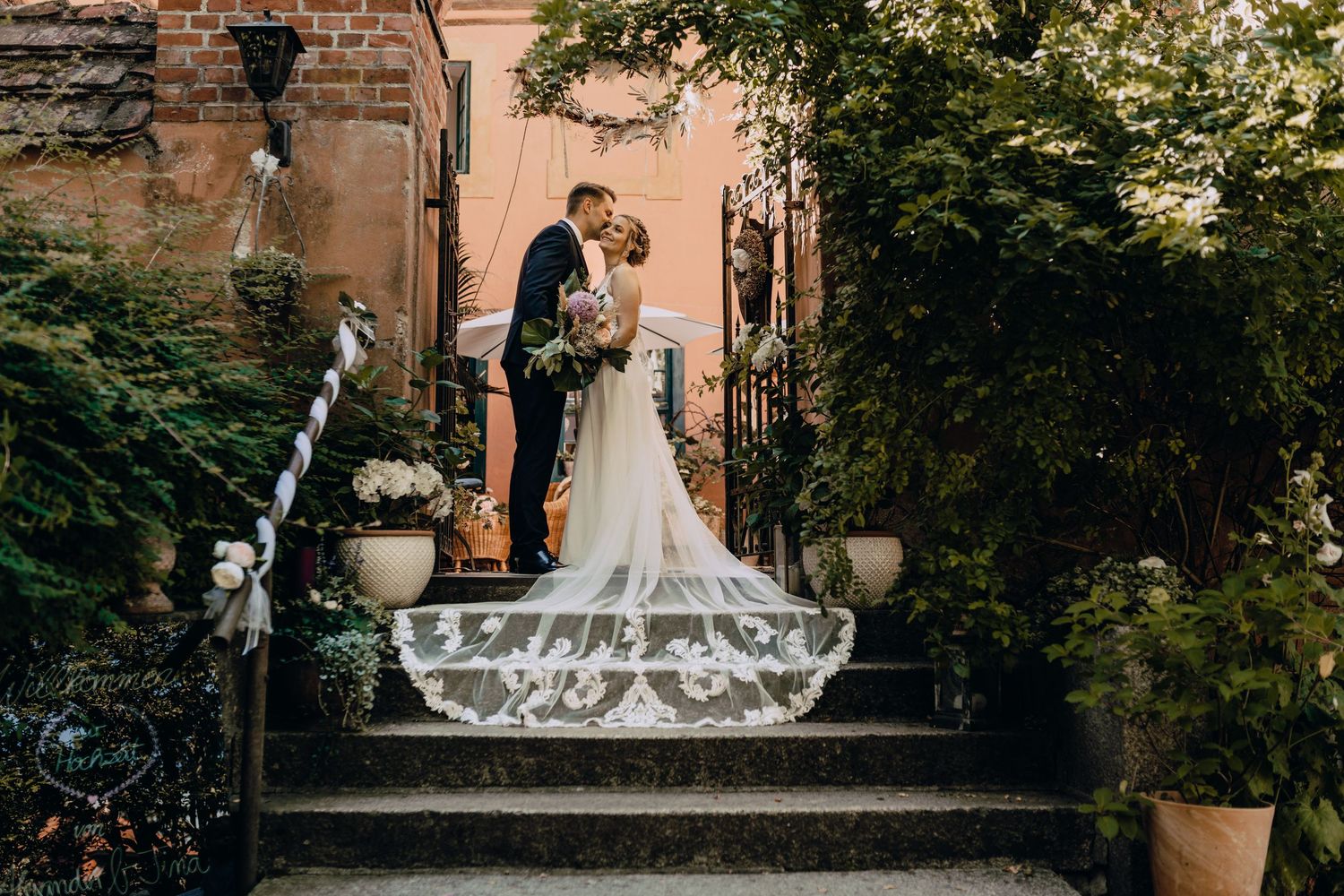 A couple shares an intimate moment on stone steps with lush greenery and brick walls surrounding them.