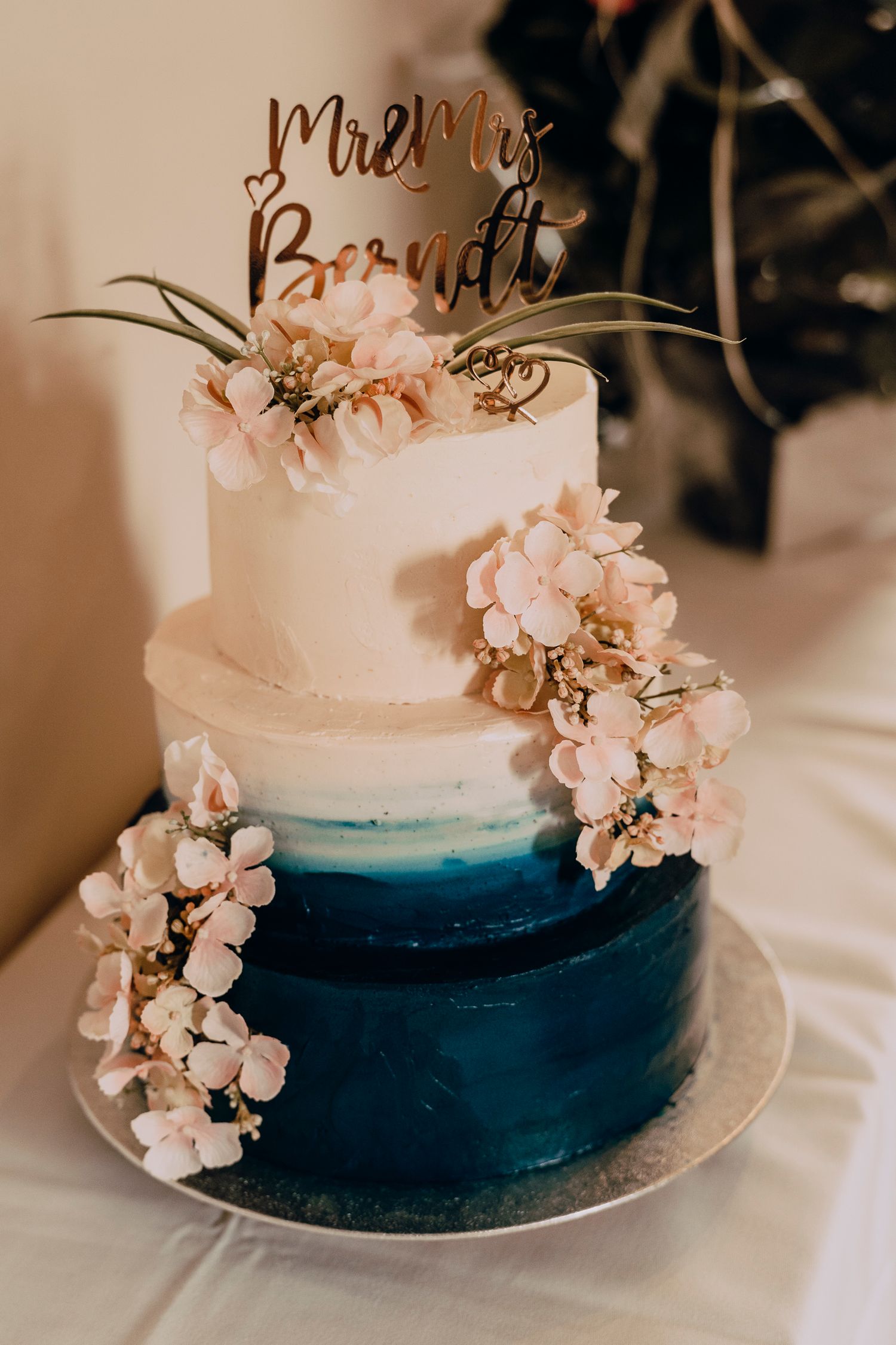 A three-tier wedding cake with white to navy blue ombre design decorated with delicate pink flowers and gold topper.