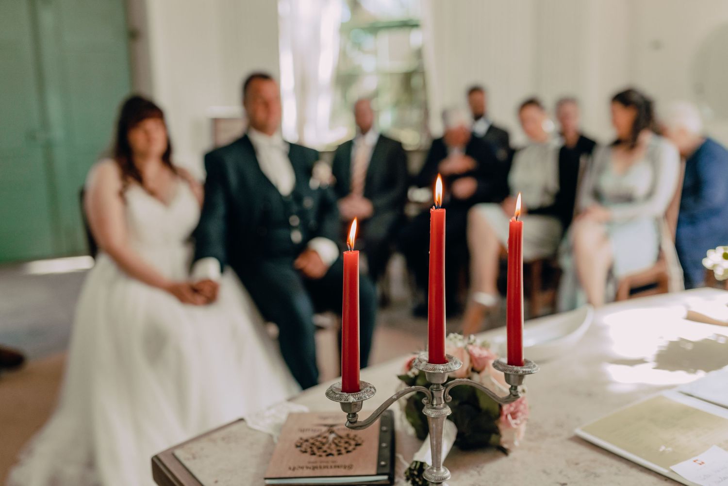 Red candles and floral decorations adorn a ceremony table with guests seated in the background.
