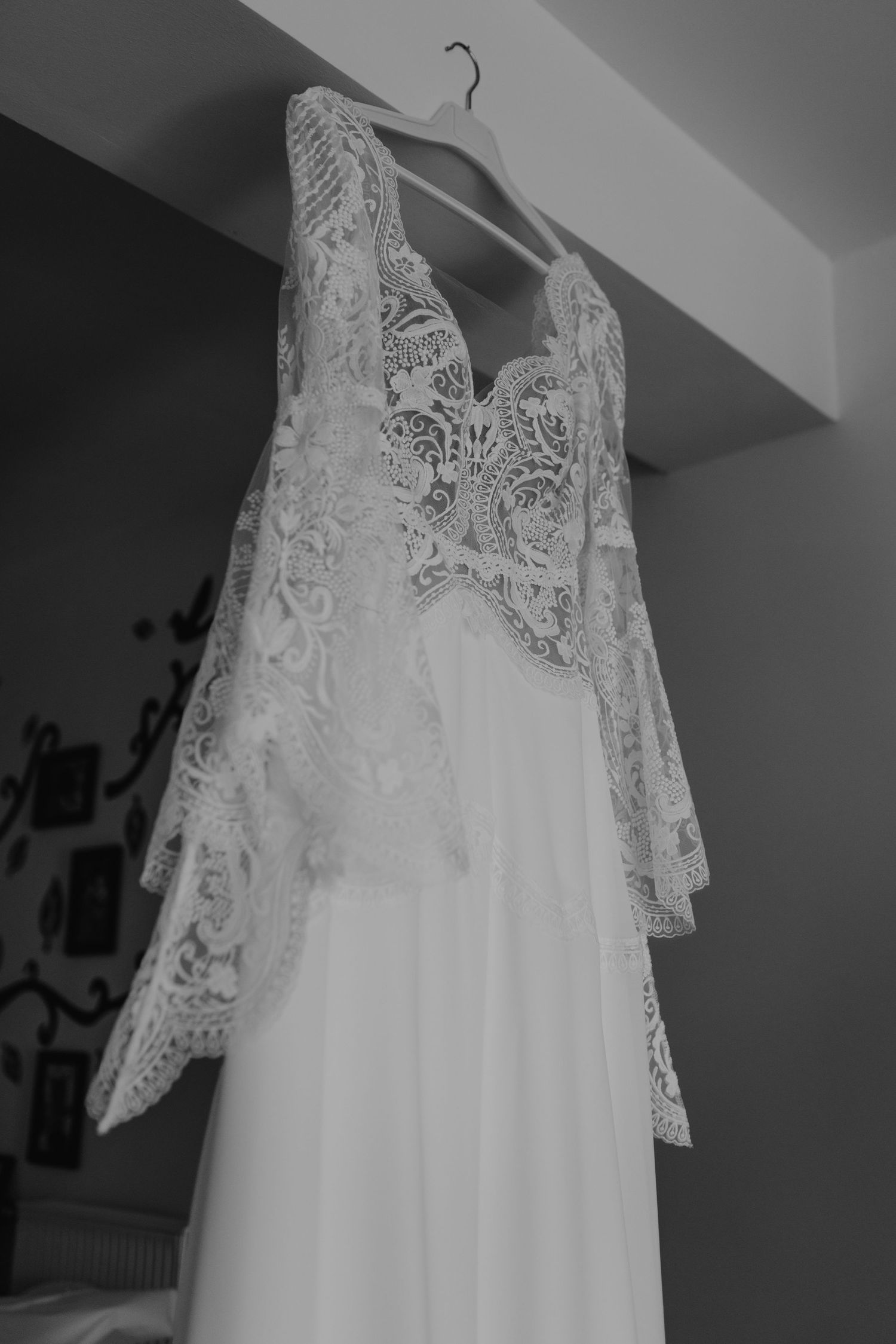 A delicate white lace wedding dress with intricate details hangs on display in black and white photography.