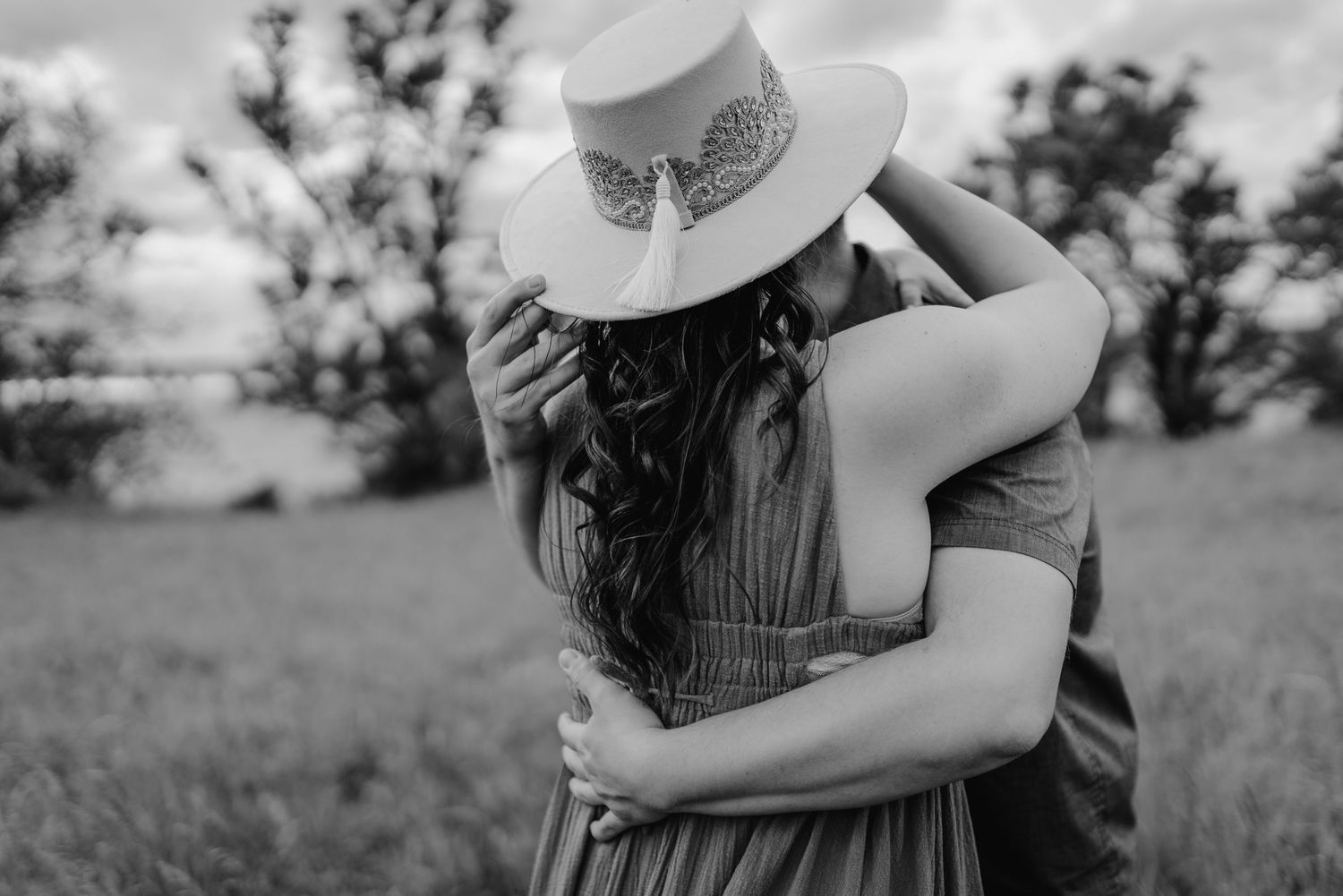 A romantic black and white photograph showing a couple embracing in a field while one wears a wide-brimmed hat.