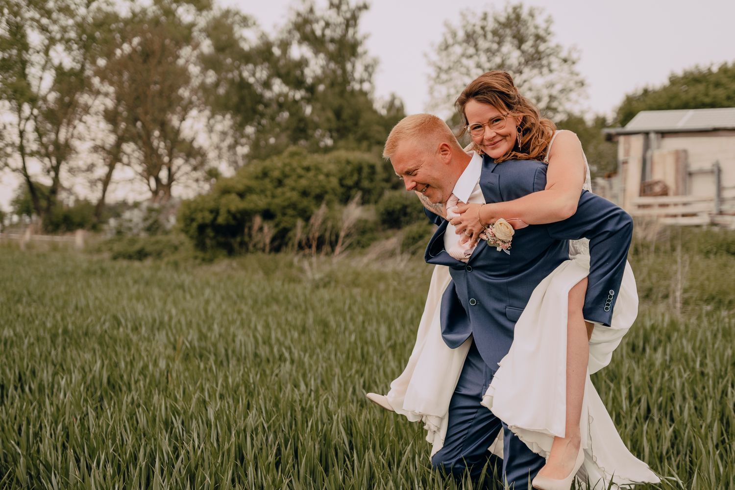 Two people share a playful moment in a grassy field, with the bride riding piggyback on her companion during their wedding.