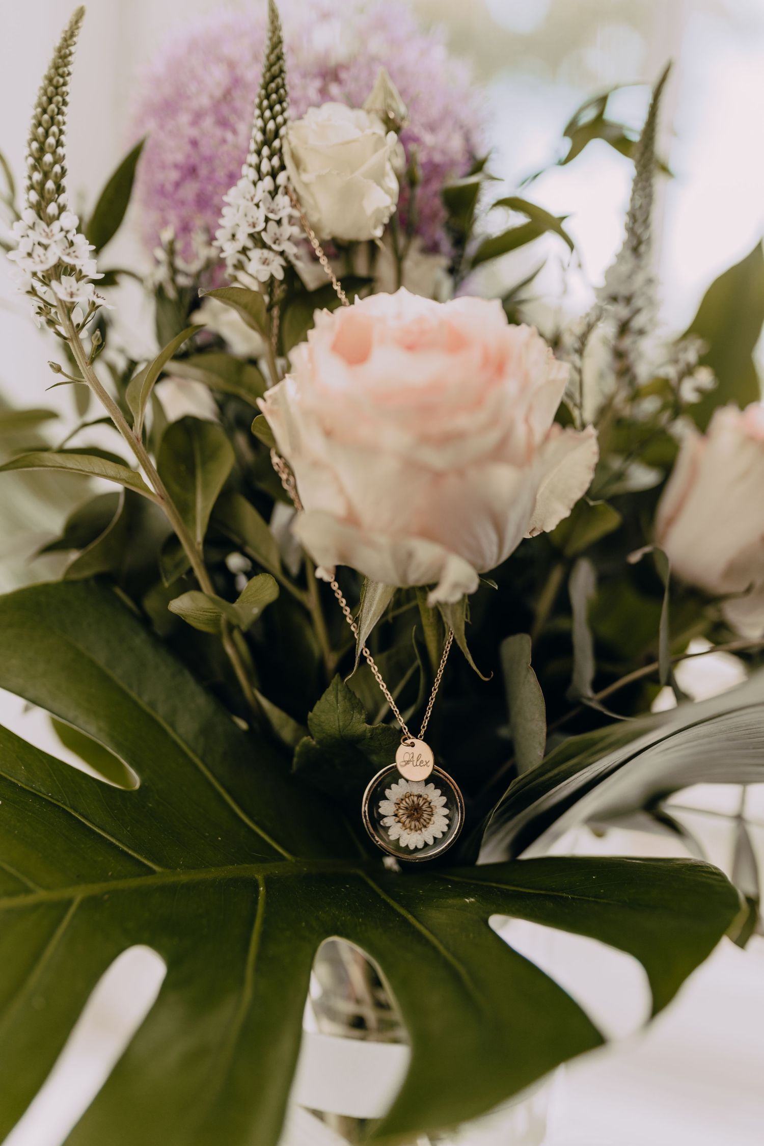 A delicate rose gold necklace pendant displayed among soft pink roses and green foliage on a white surface.