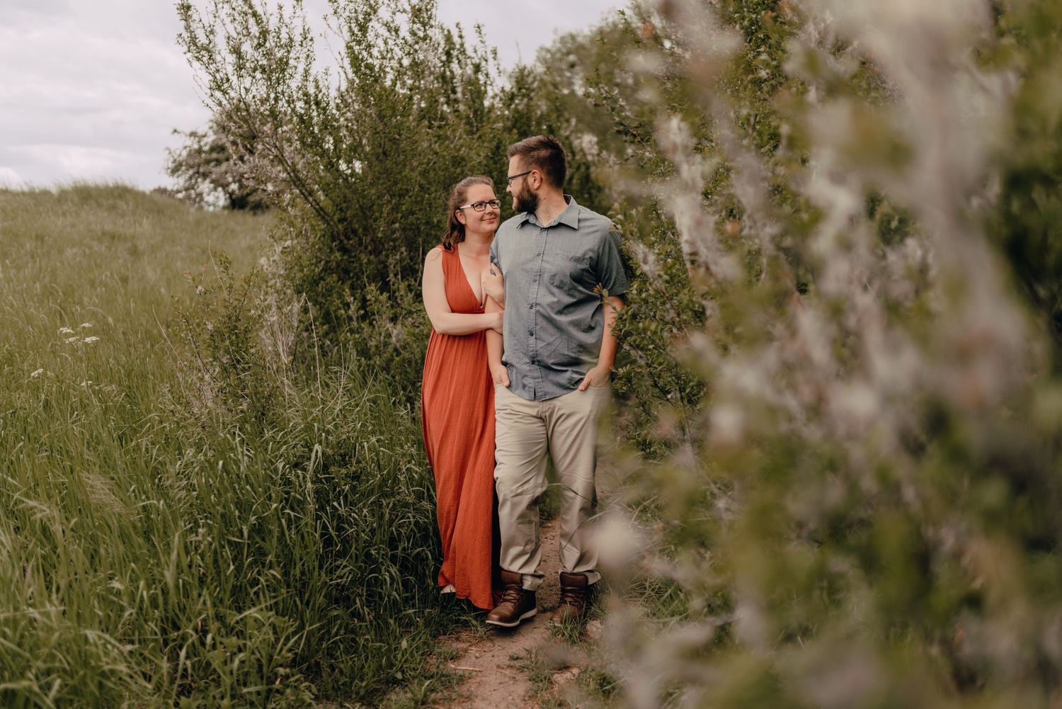 A couple stands together on a grassy path, with her in a long orange dress and him in casual attire with greenery around them.