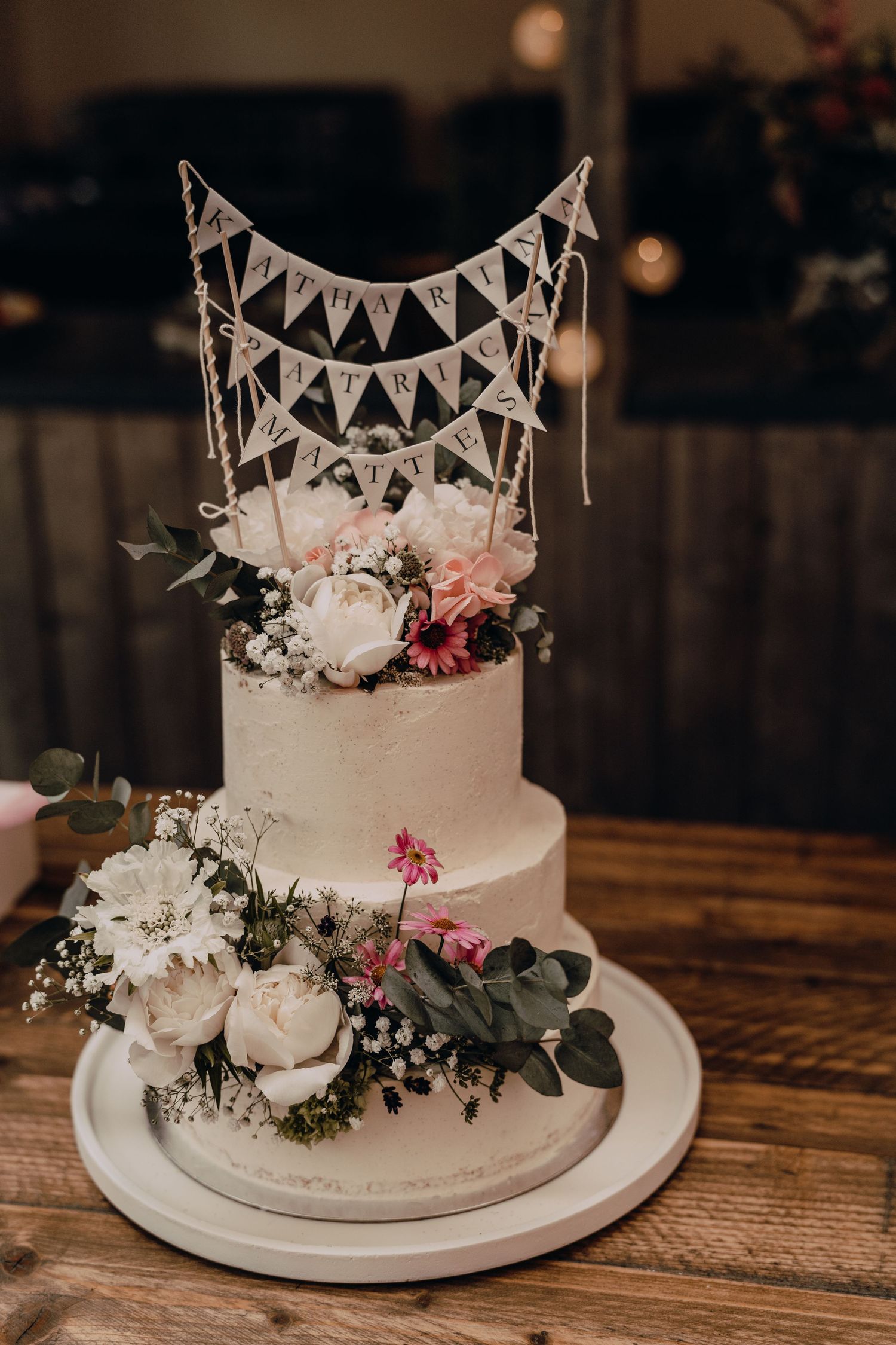 Elegant white two-tier wedding cake decorated with pink flowers, eucalyptus leaves and bunting topper on wooden table.