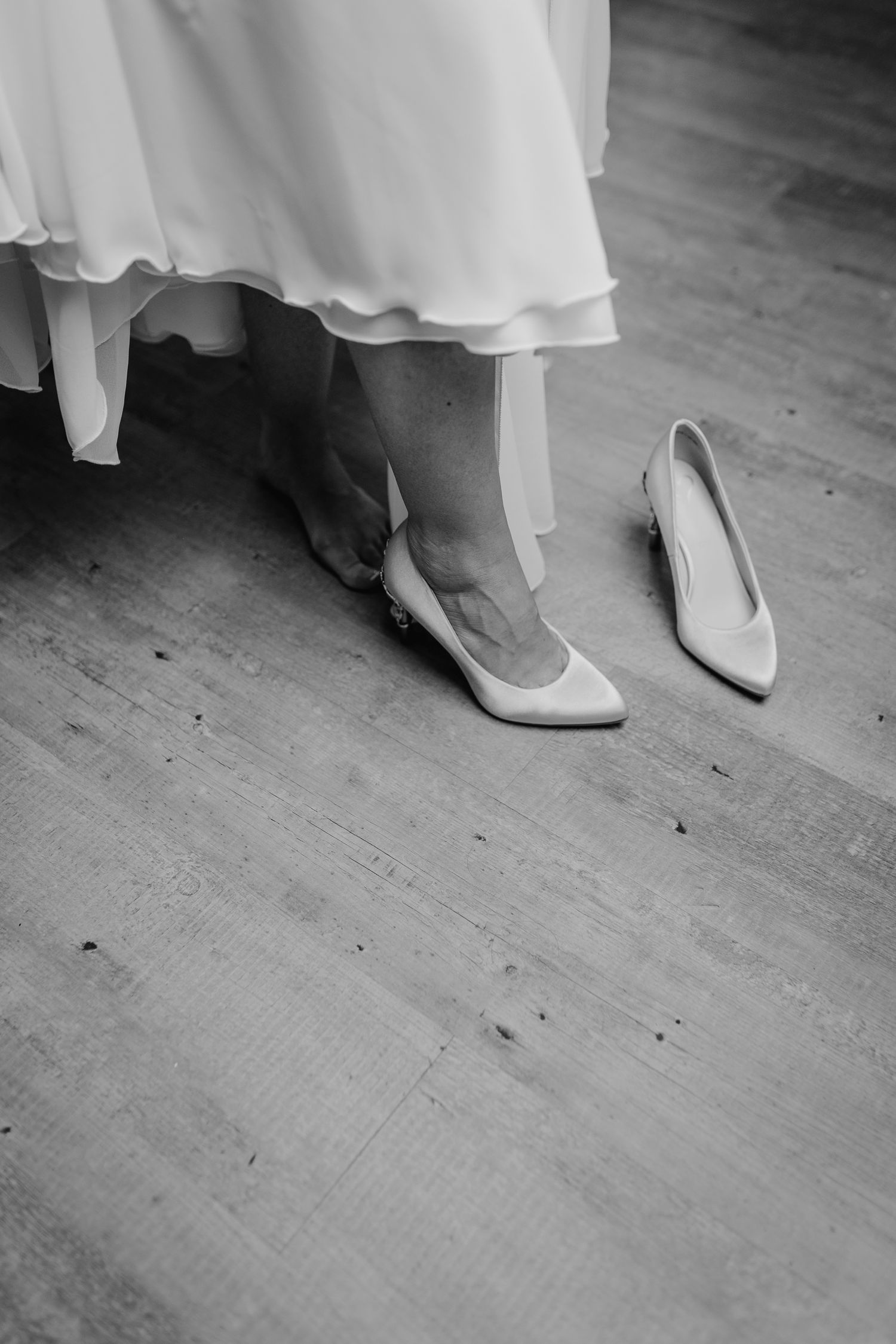 Black and white photo showing elegant white high heels on a wooden floor with flowing white fabric above.
