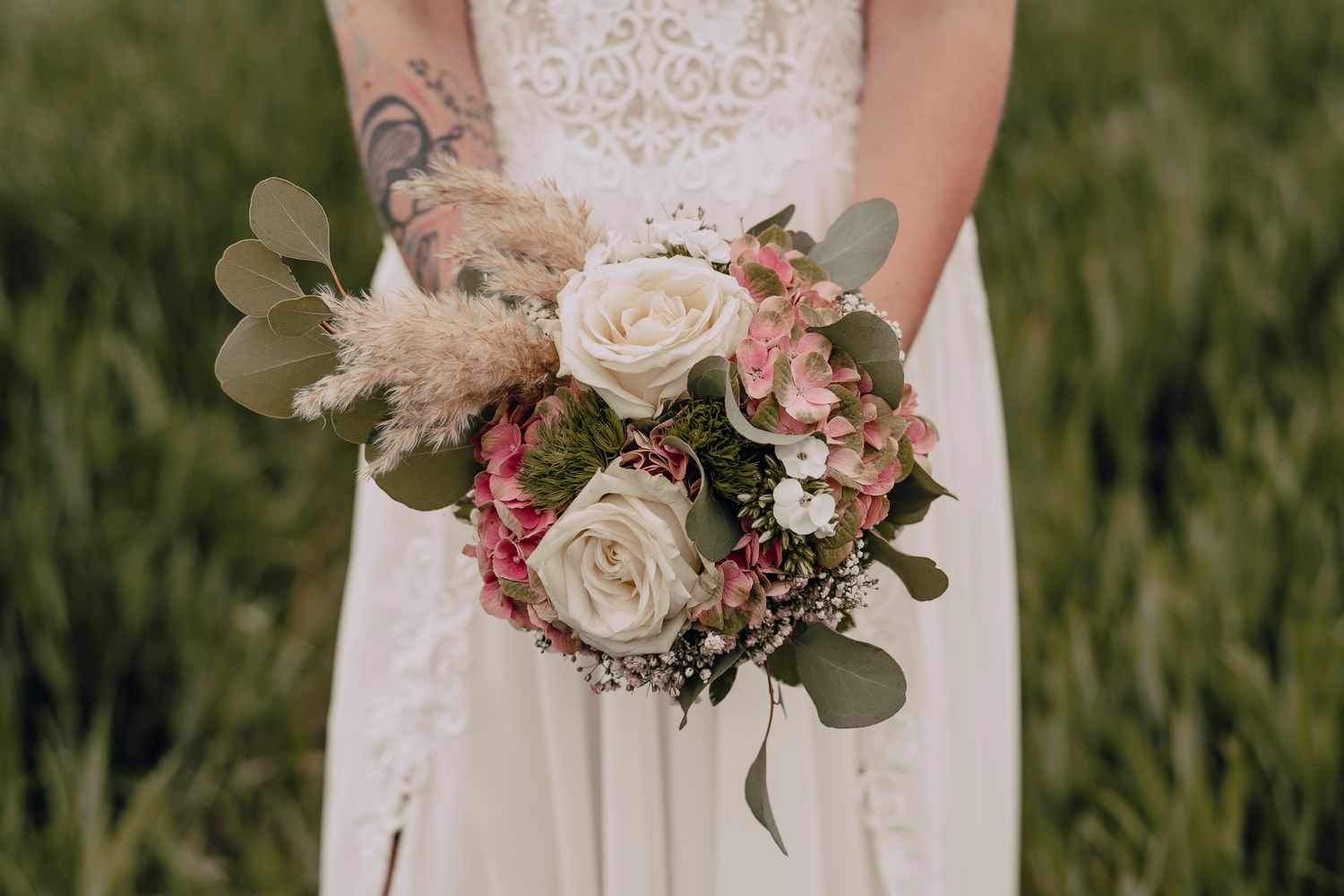 A vintage-style wedding bouquet with cream roses, pink accents, and eucalyptus leaves held against a white lace dress.