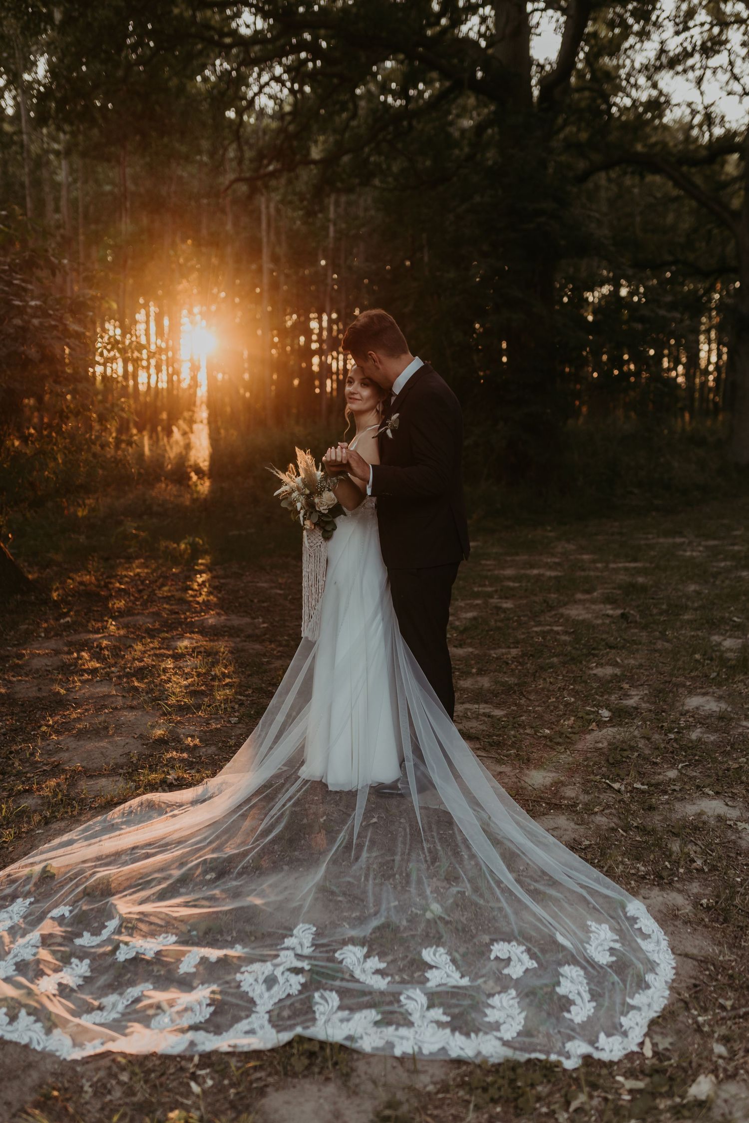 Silhouetted couple shares romantic moment at sunset with bride's long lace veil trailing behind on forest floor.