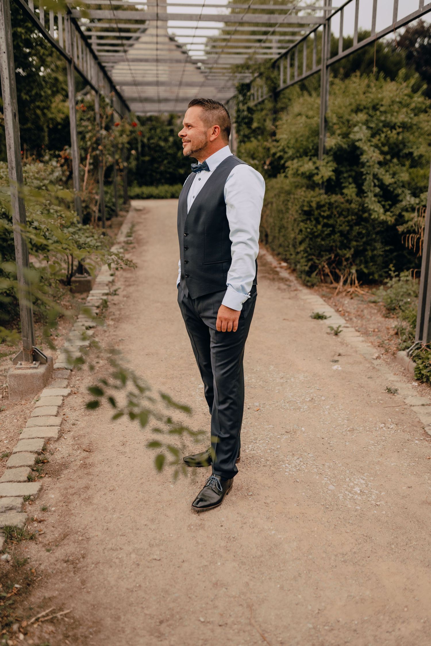 Side view of a groom in formal attire standing in a garden walkway with an overhead pergola structure.