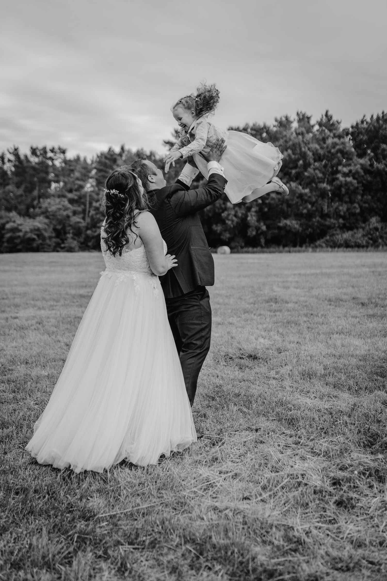Wedding couple embracing in a black and white photo on a grassy field with pine trees in background.