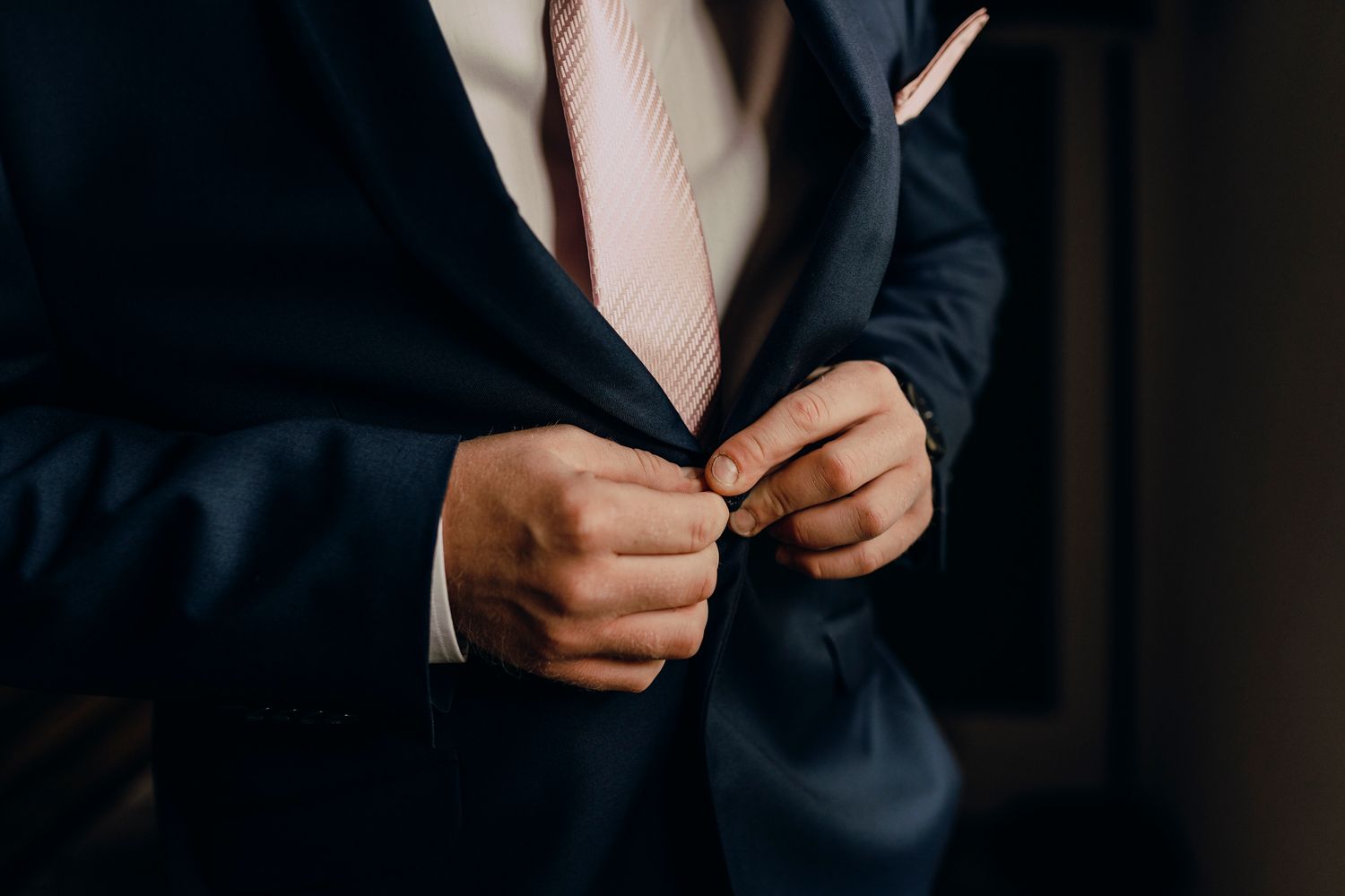 Close-up view of hands buttoning a navy blue suit jacket with white dress shirt and pink tie visible.