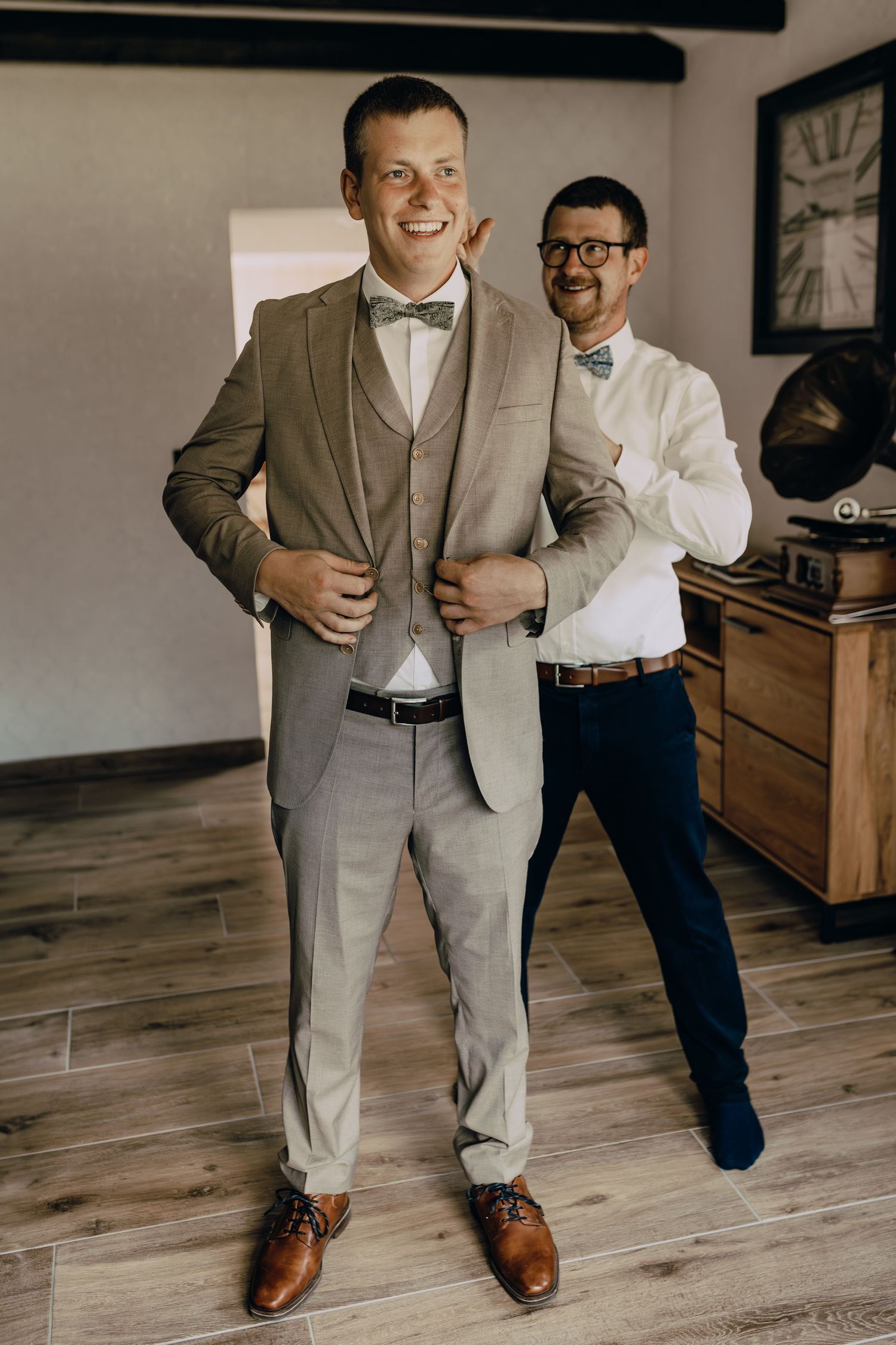Two people in wedding attire with bow ties stand on hardwood floor in room with vintage decor.