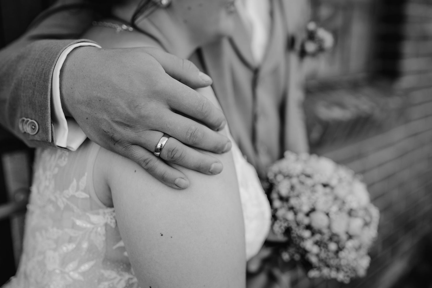 Black and white close-up of loving embrace during wedding ceremony with bridal bouquet in background.