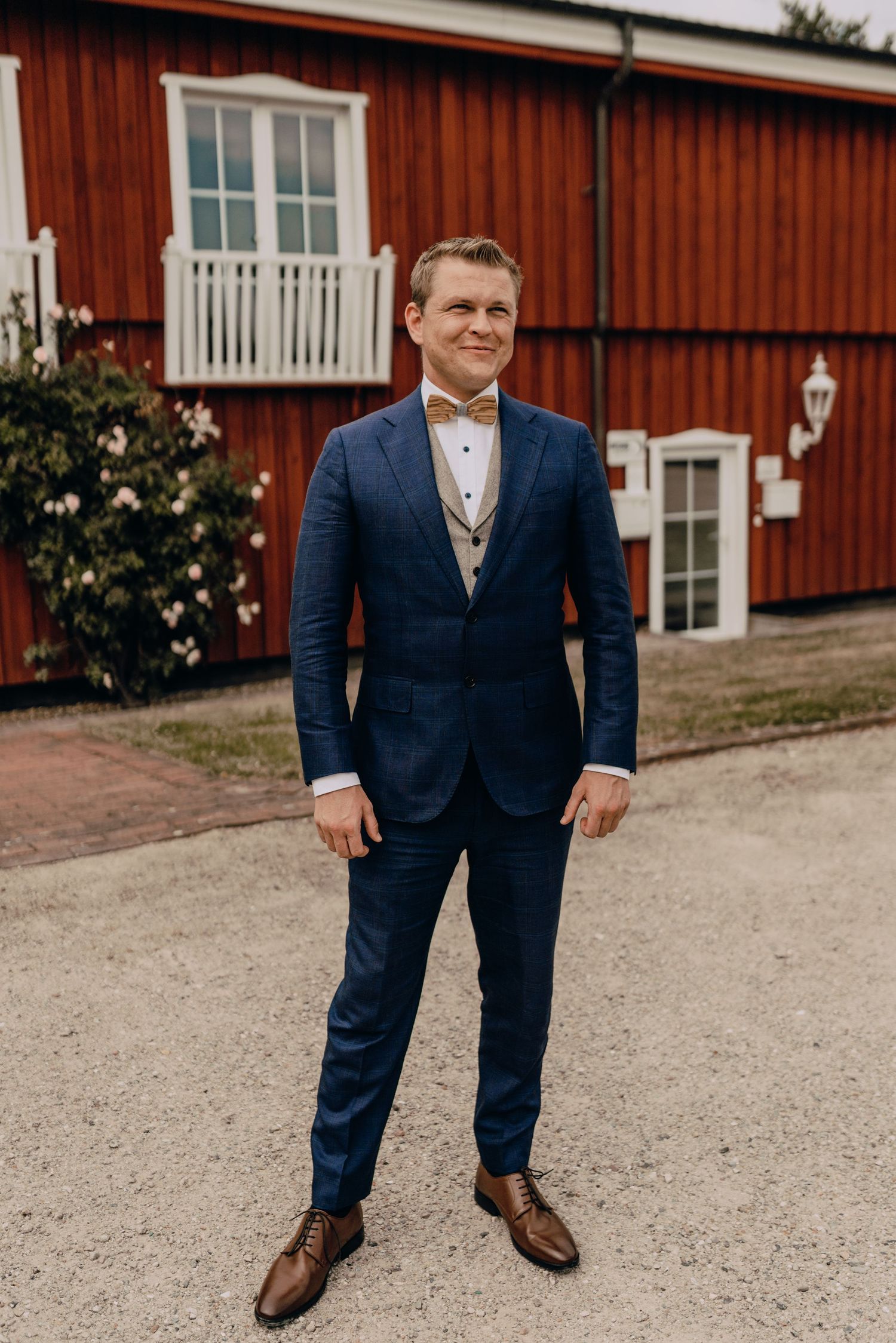 Groom wearing navy blue suit and bow tie stands in front of red barn building.