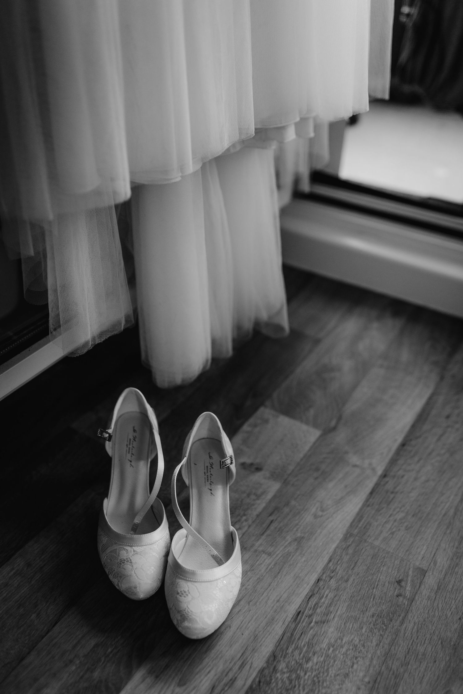 White wedding dress hanging above elegant bridal shoes on a wooden floor.