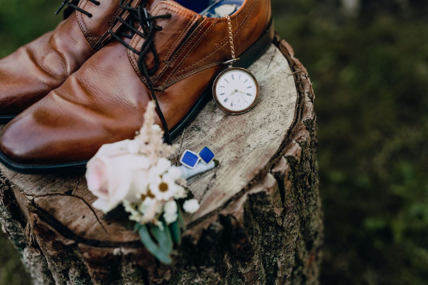 Brown leather dress shoes with vintage pocket watch and delicate white rose arranged on rustic wood stump.