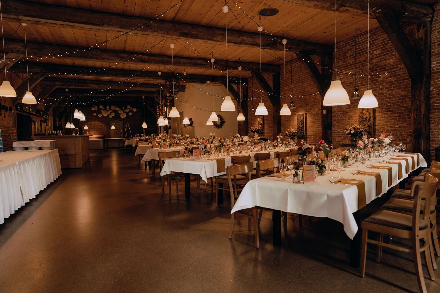 Rustic wedding reception venue with long tables and white pendant lighting.
