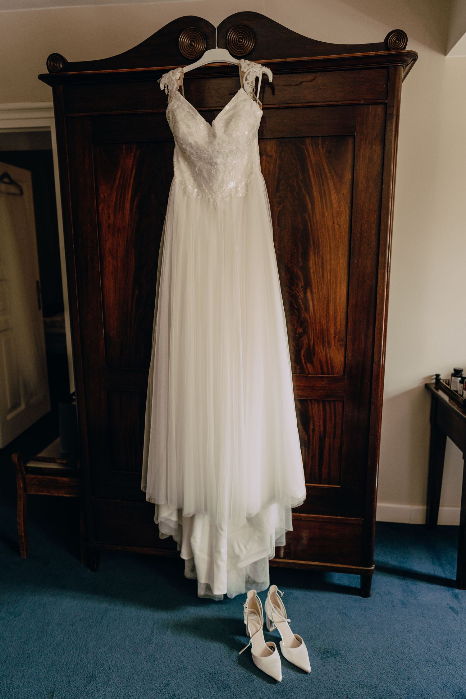 An elegant white wedding dress hangs from a dark wooden armoire with matching white heels placed beneath it.