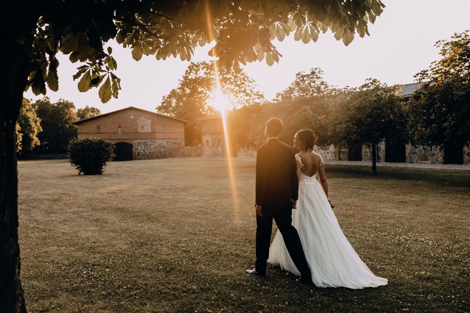 Silhouetted couple in wedding attire walking at sunset on expansive lawn.