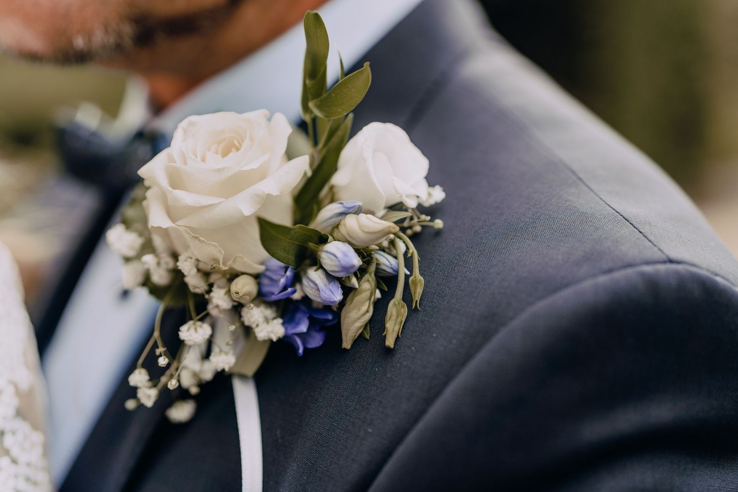 A delicate white rose boutonniere with blue flowers and baby's breath pinned to a navy suit lapel.