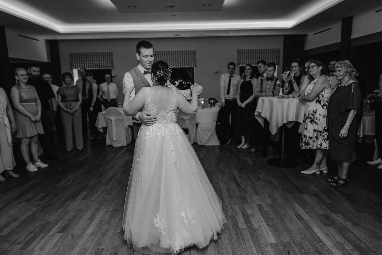 A couple shares their first dance on a wooden floor at their wedding reception while guests watch from the sidelines.