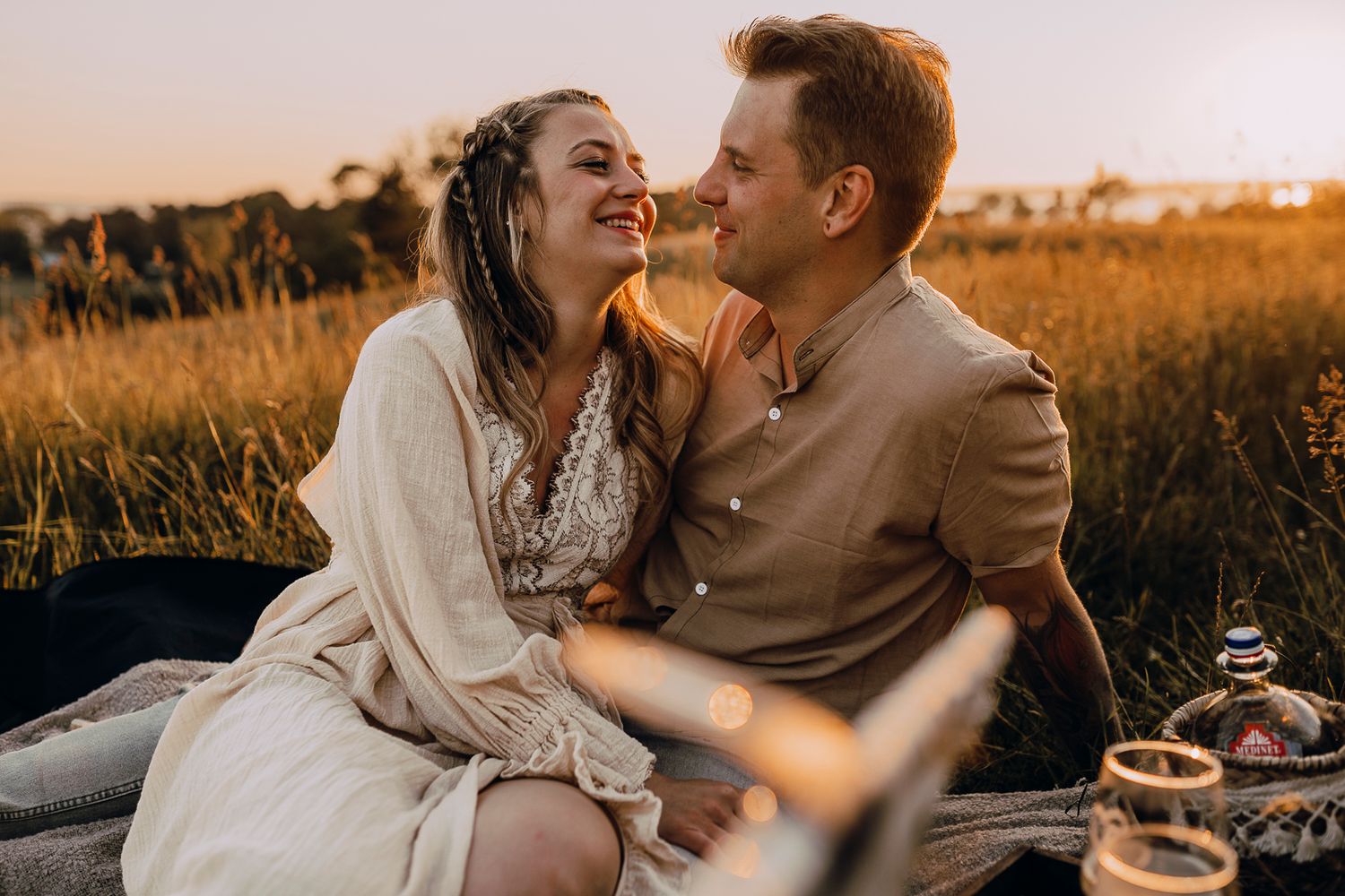 A couple shares an intimate moment during a sunset picnic in a golden field wearing light colored casual attire.