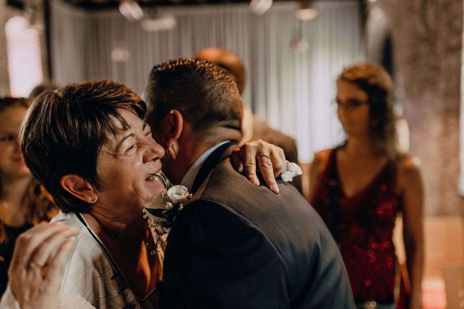 Two people share an emotional embrace at a wedding reception while a guest in red watches in the background.