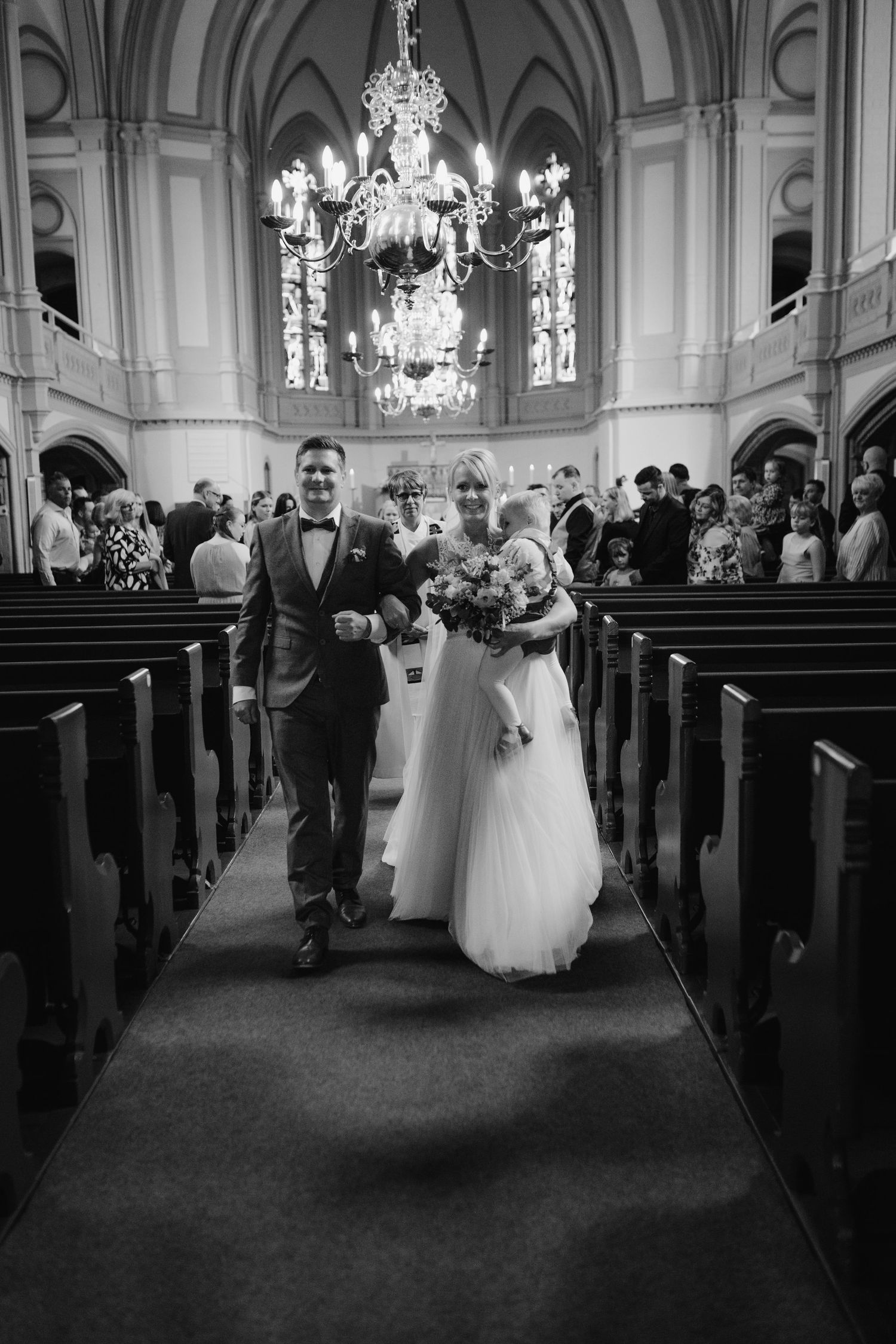 A wedding couple walking down the aisle of a grand church with ornate chandeliers and stained glass windows.