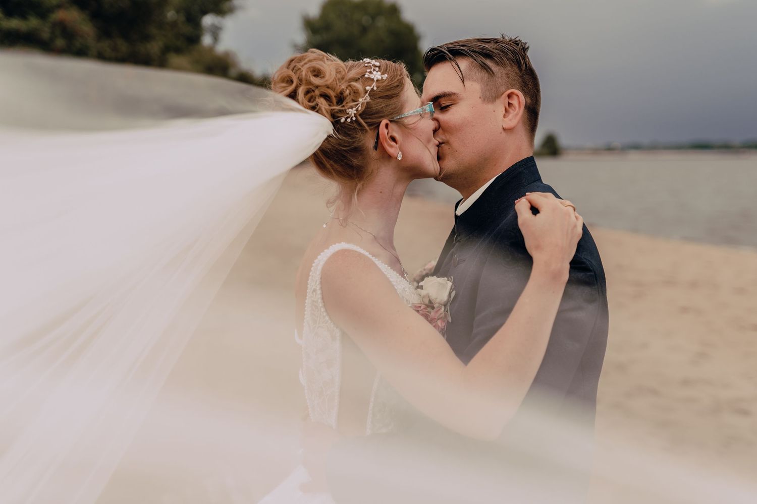 A romantic wedding couple share a tender kiss on a beach as the bride's veil flows in the wind.