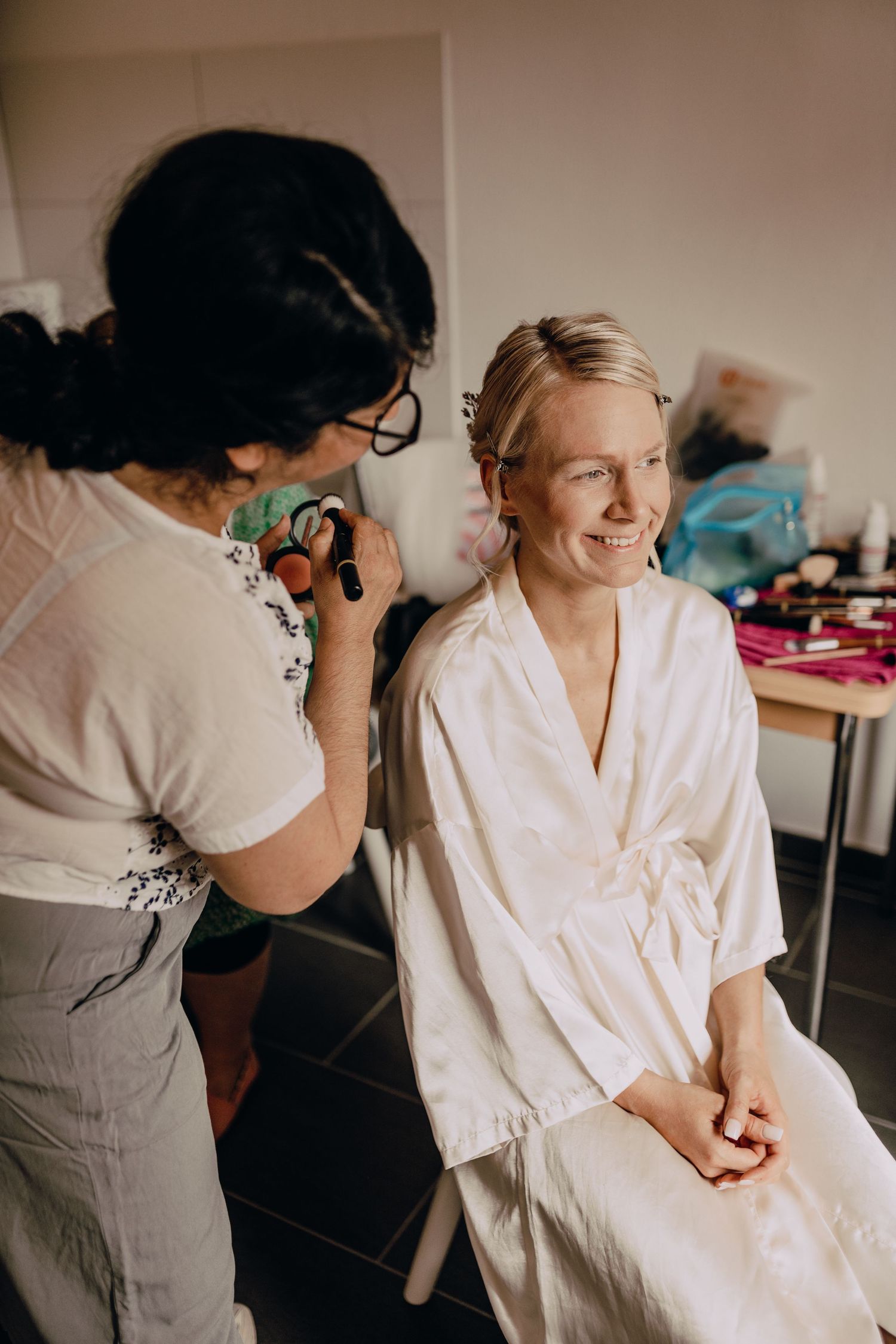 Makeup artist applies cosmetics to a client wearing a white robe in a getting-ready room.