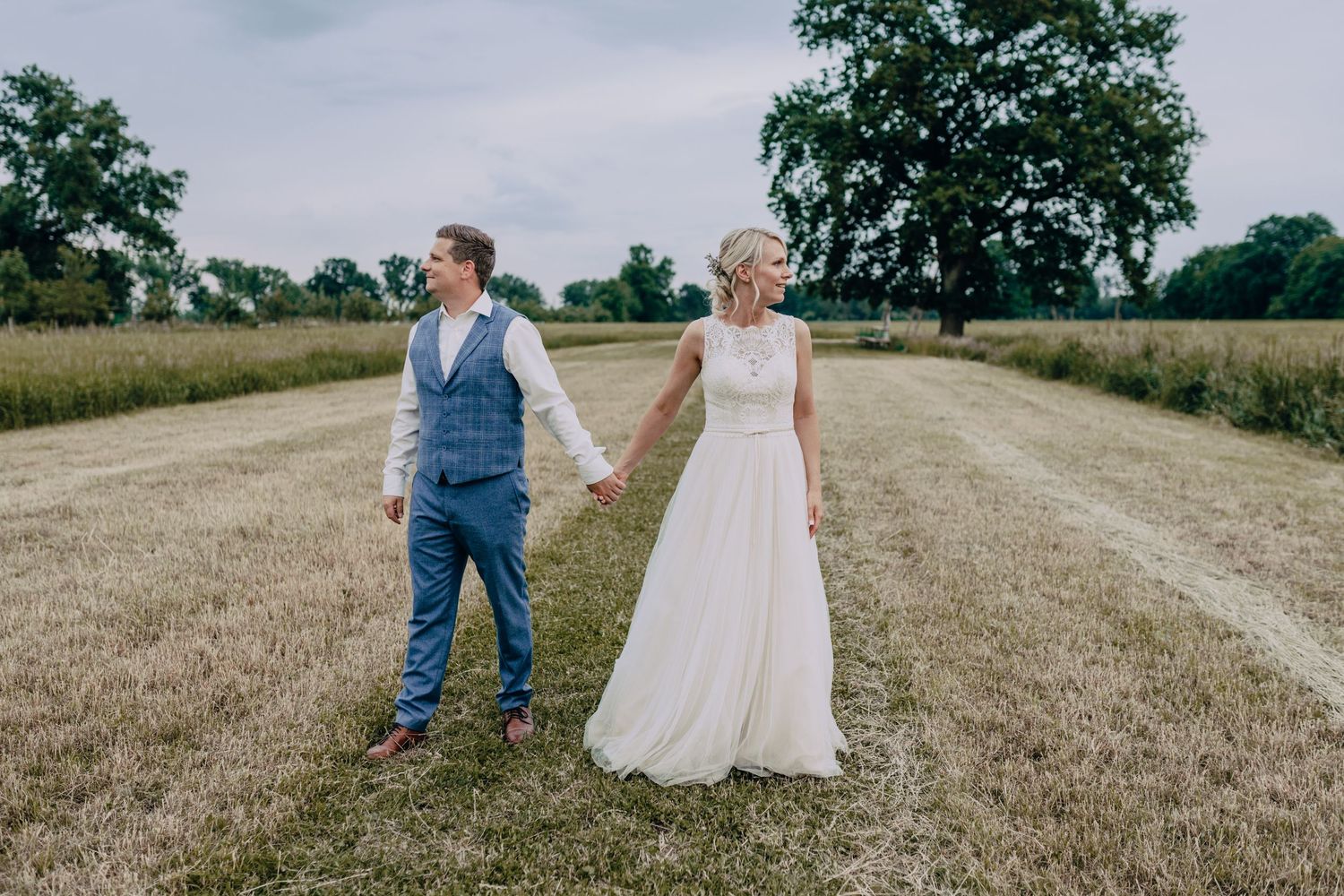 A couple holds hands while standing back-to-back on a dirt path between rural fields on their wedding day.