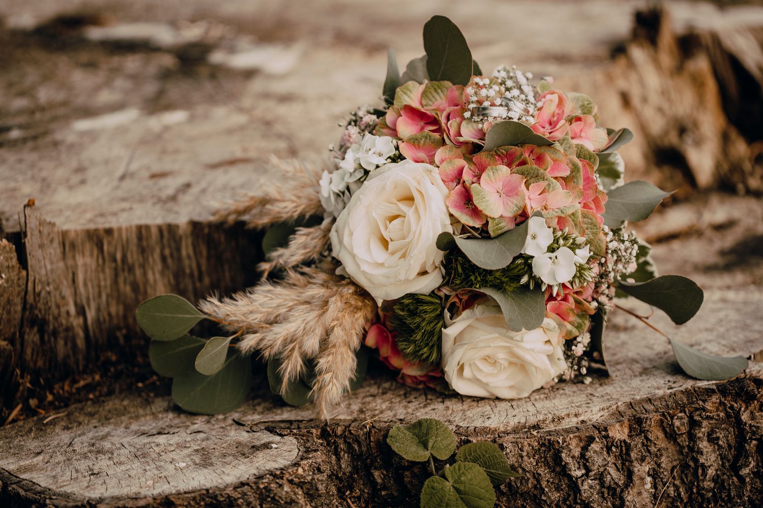 Wedding bouquet with white roses, pink blooms, pampas grass and eucalyptus leaves rests on rustic wood stump.