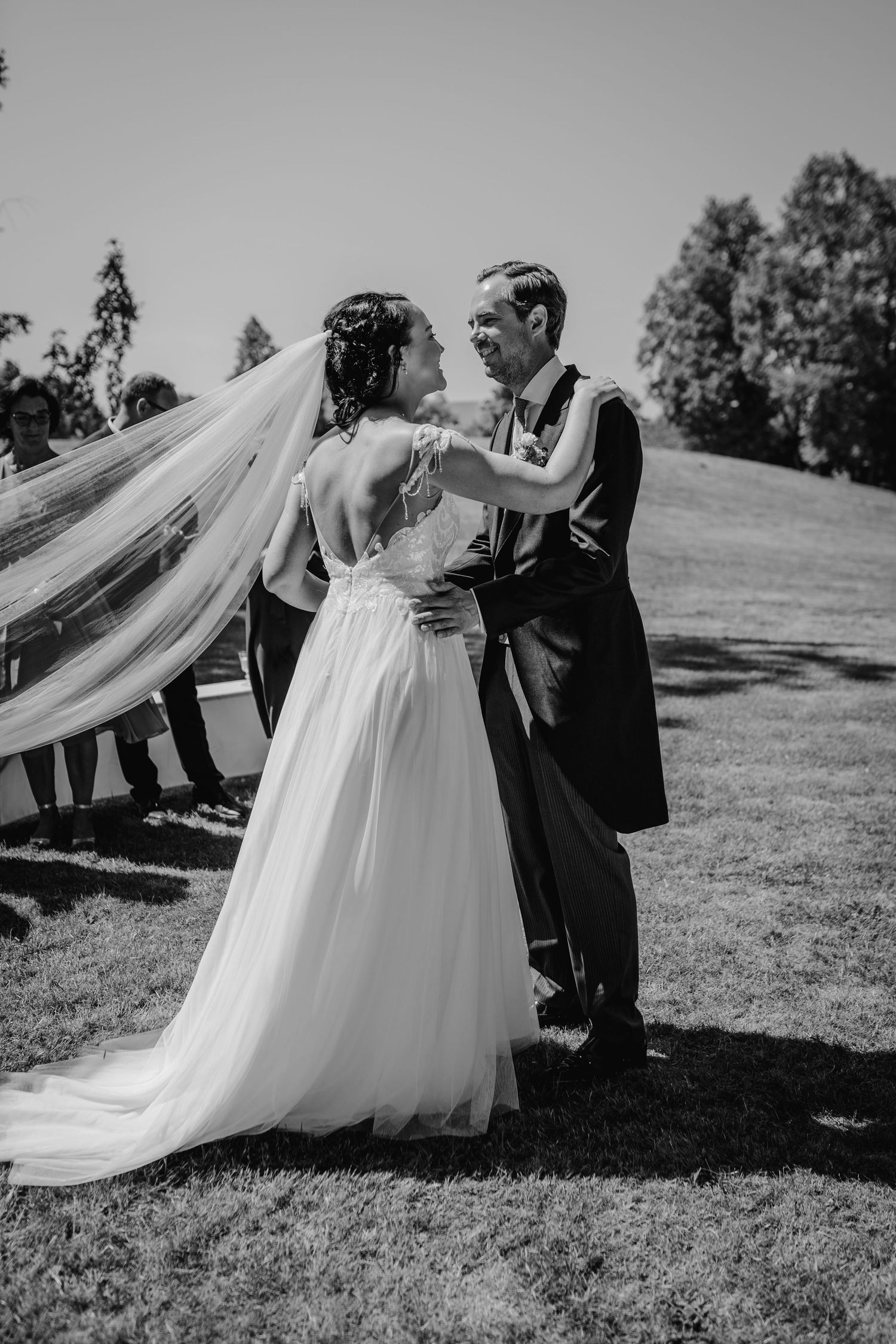 Newlyweds share intimate moment on sunny lawn with flowing bridal veil.