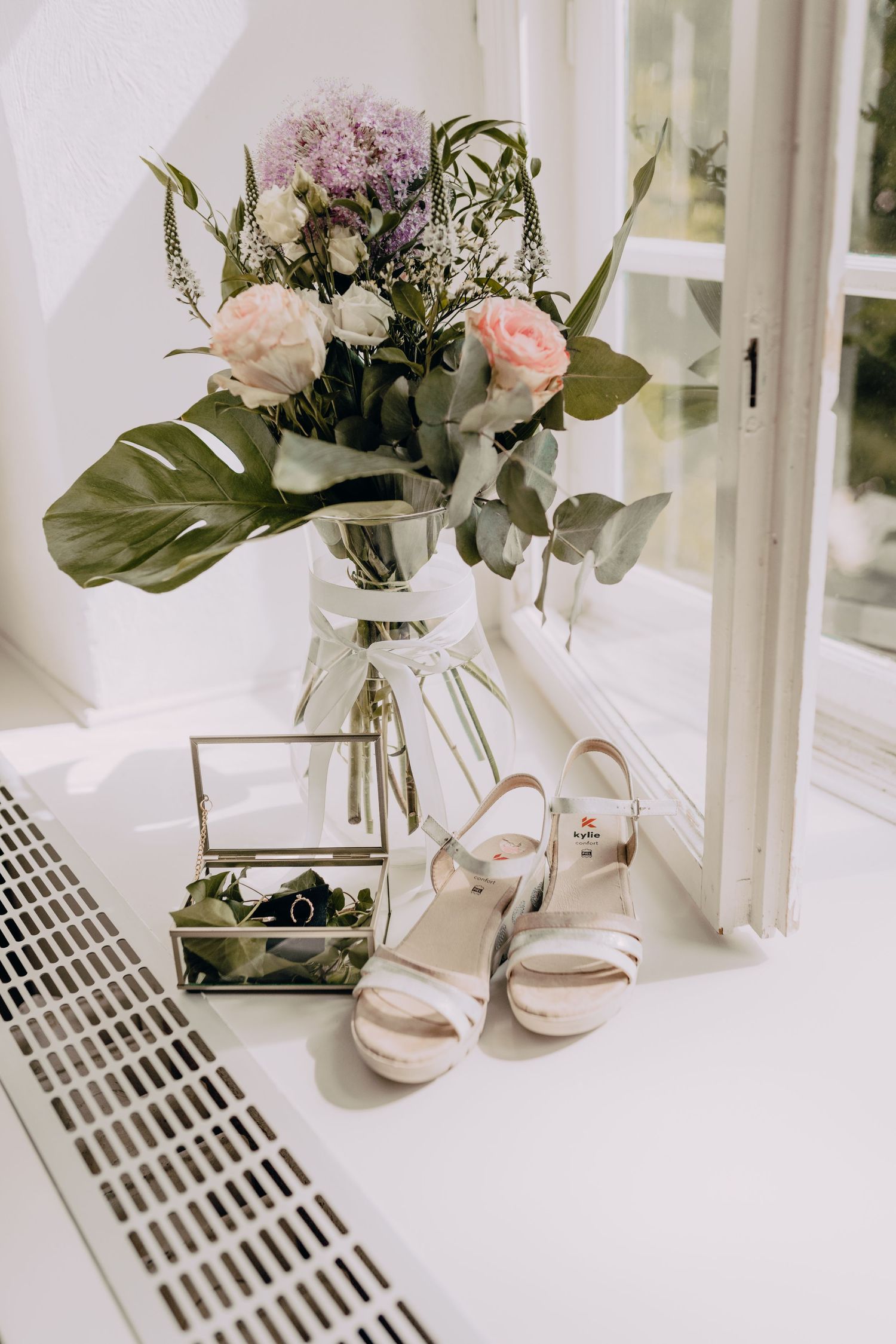 Elegant white and pink floral arrangements in glass vases next to delicate bridal sandals on a bright windowsill.
