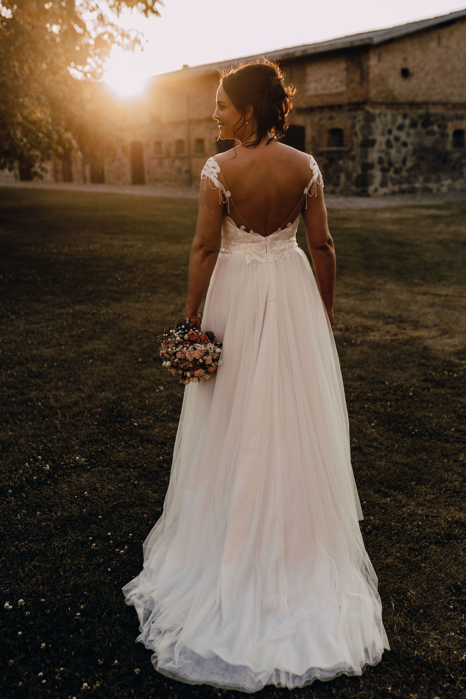 Bride in flowing white gown with open back design holding bouquet at sunset.