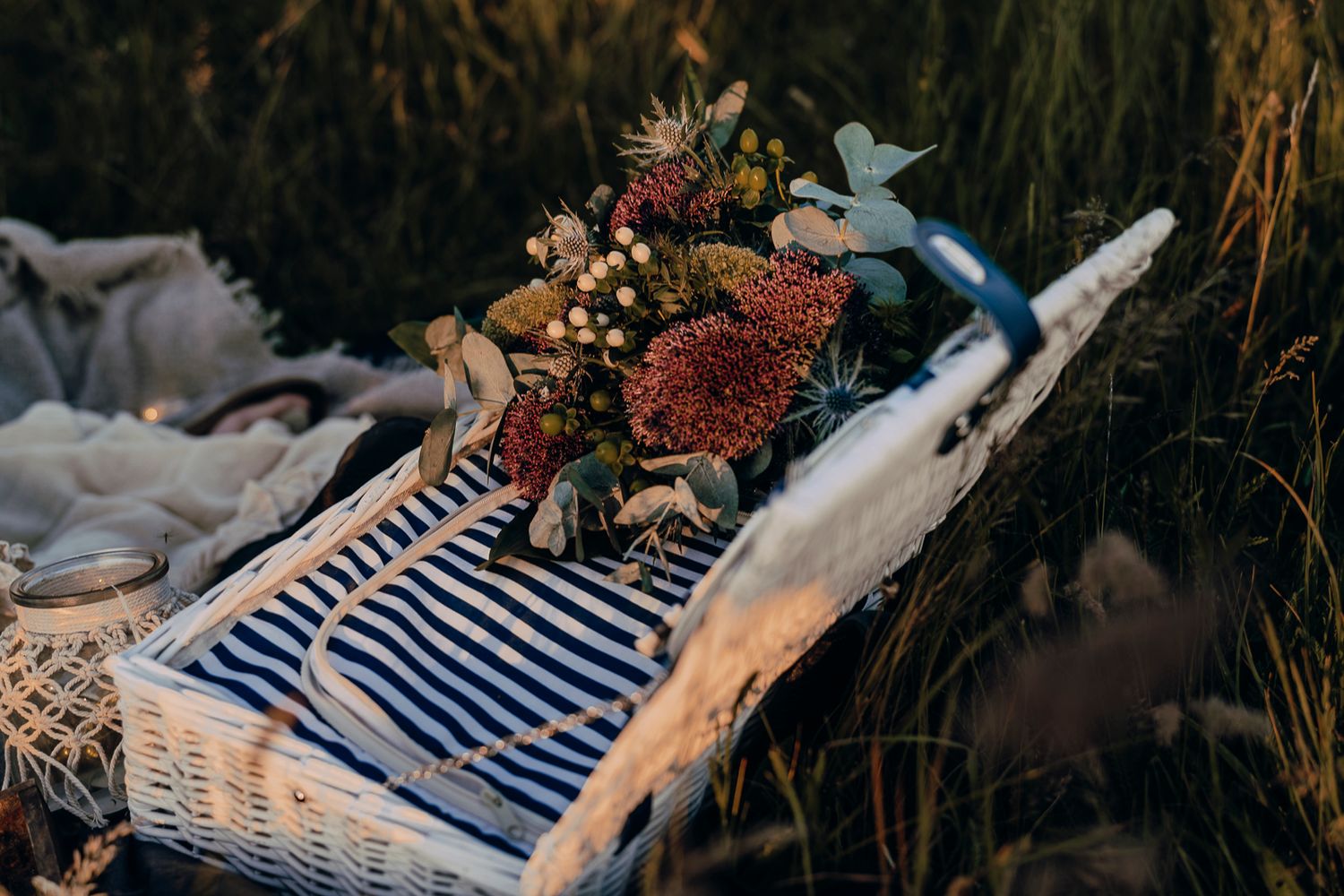 A vintage white wicker picnic basket with blue striped lining and autumn flowers rests in tall grass at sunset.