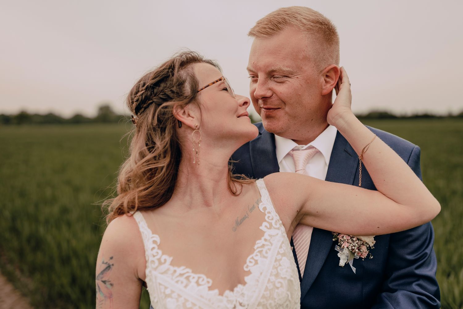 A couple in wedding attire embraces lovingly in a grassy field at sunset.