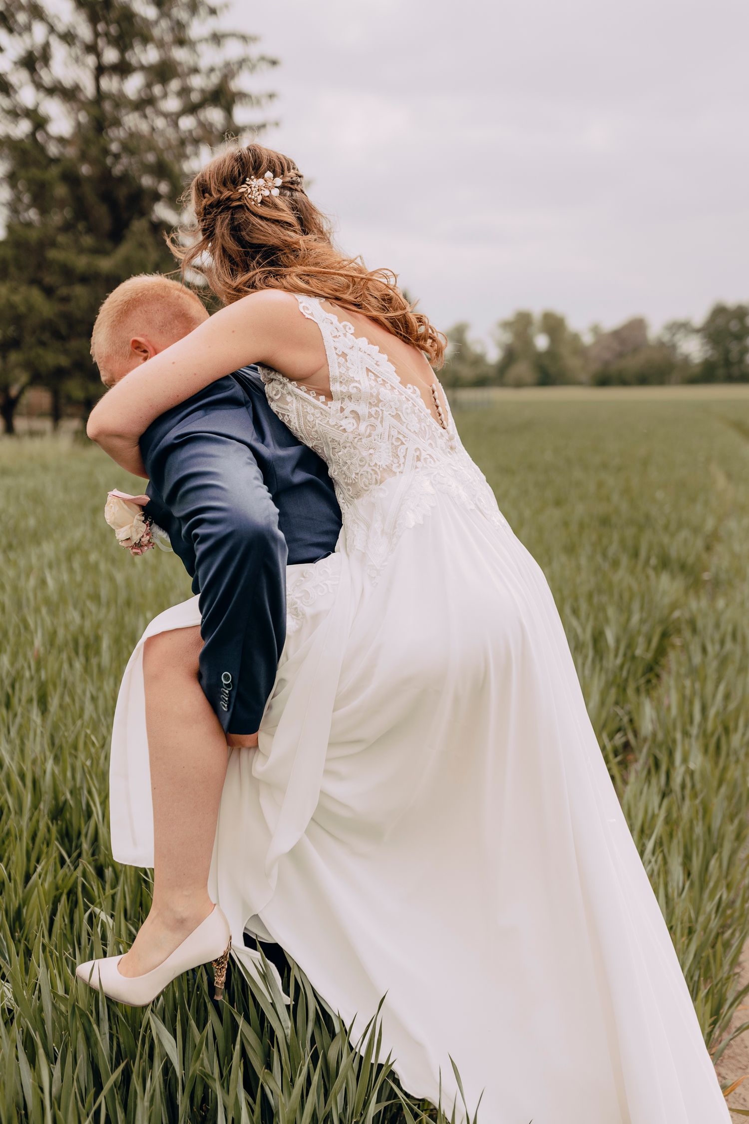 A couple embraces in a golden wheat field, with the bride wearing a flowing white lace dress and groom in navy suit.