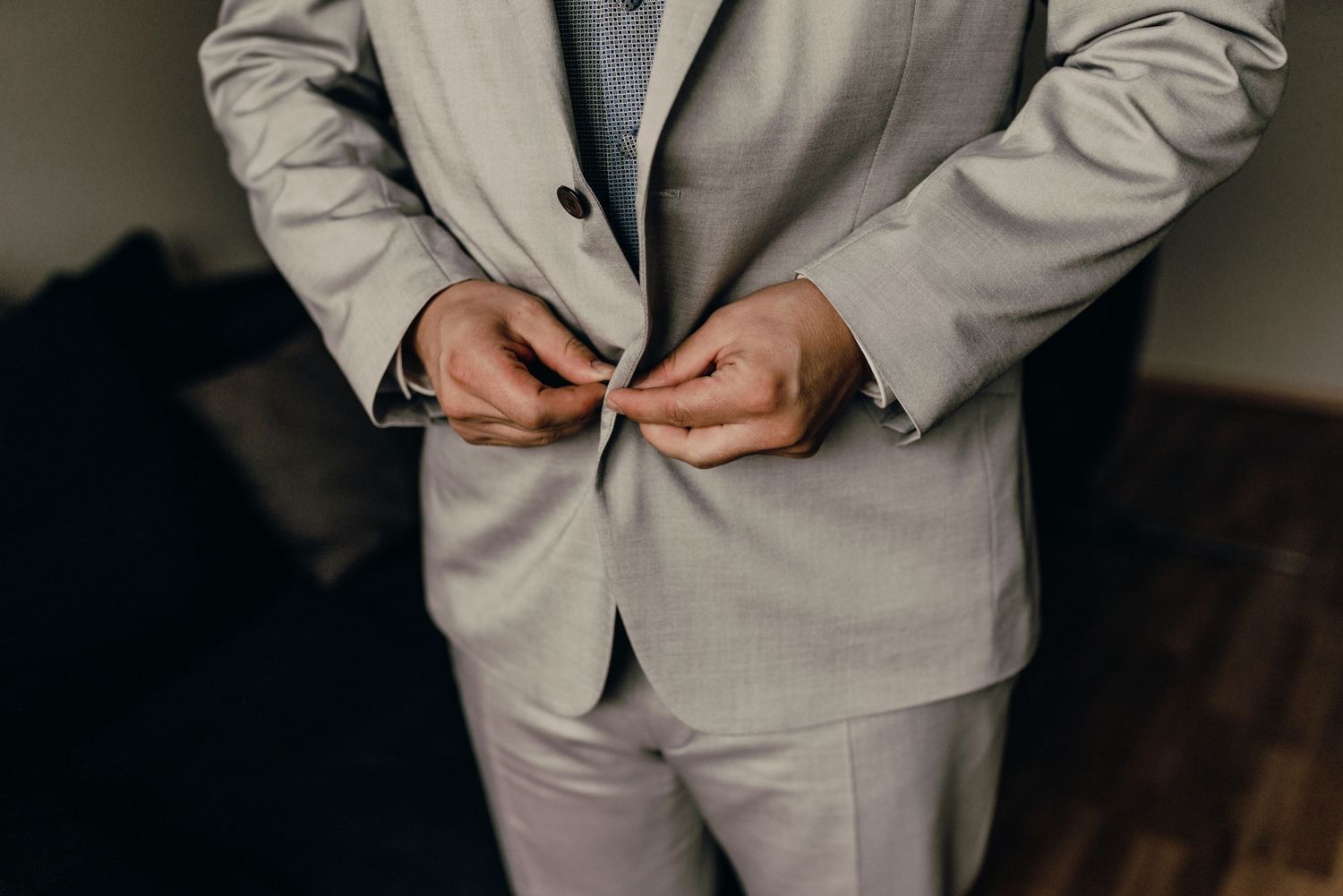 Person in a light gray suit adjusting their jacket button in a dimly lit room.