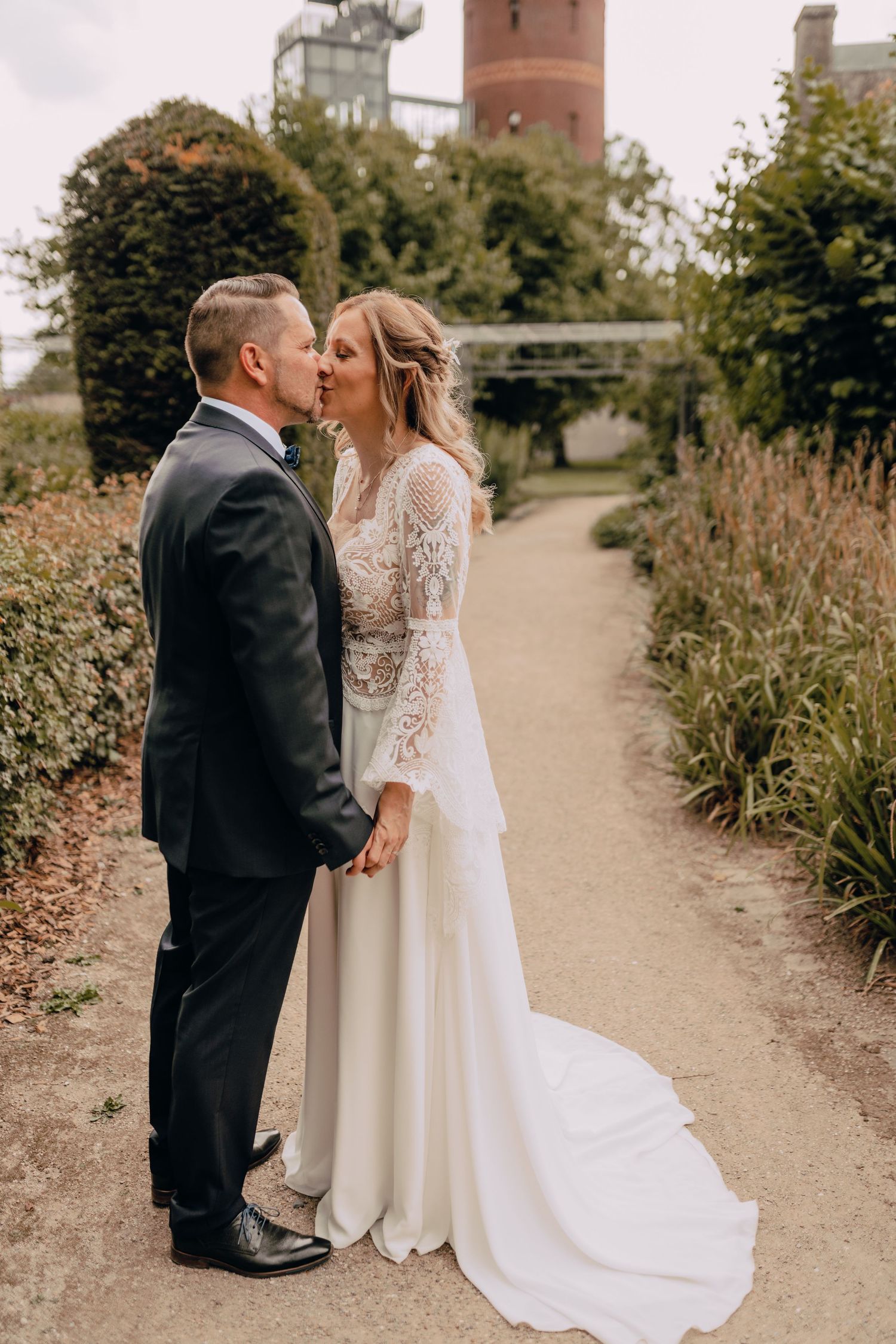 Newlywed couple shares romantic kiss on garden path with long lace sleeve wedding gown and classic black tuxedo.