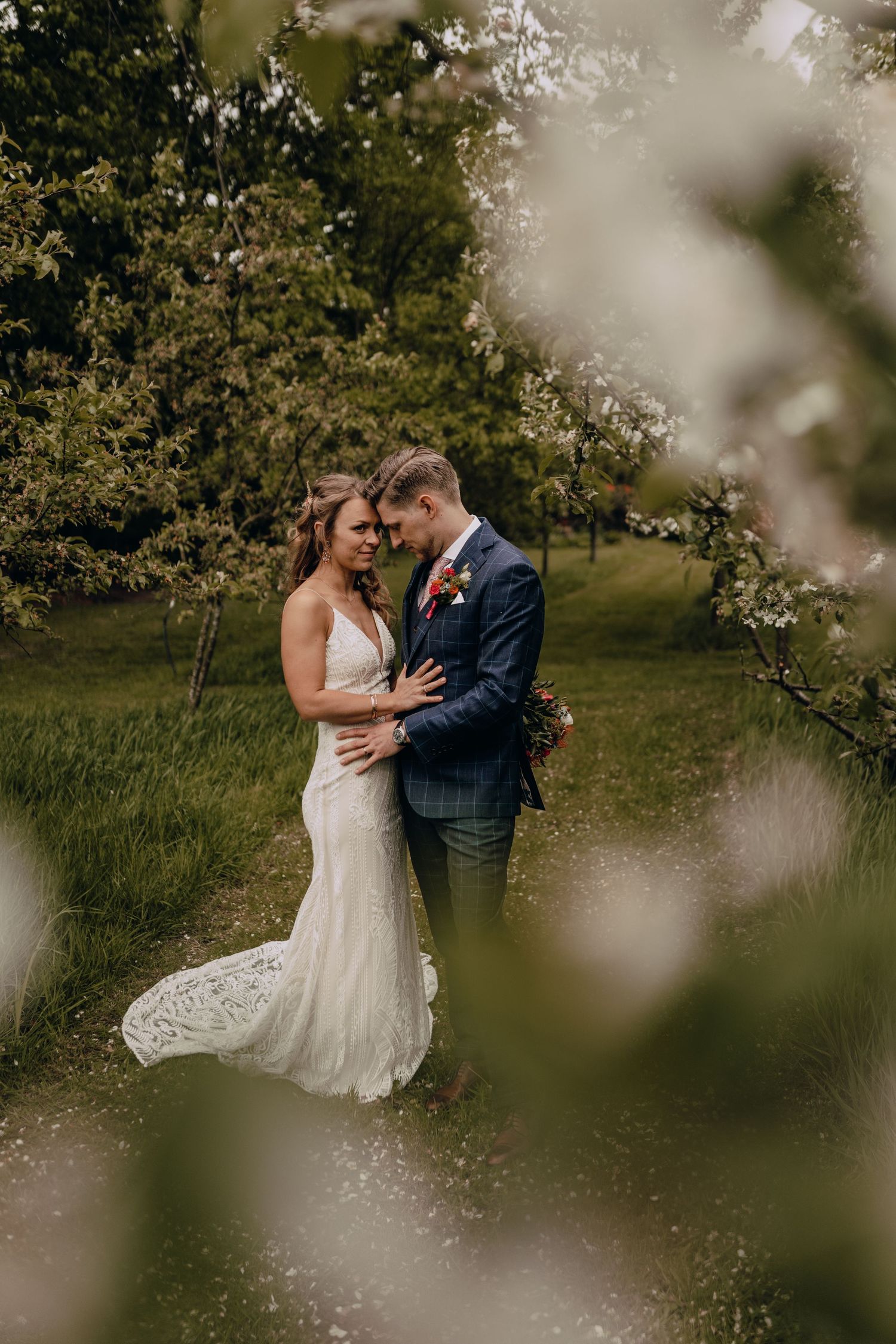 Intimate wedding portrait through blurred foliage showing couple in elegant attire embracing outdoors.