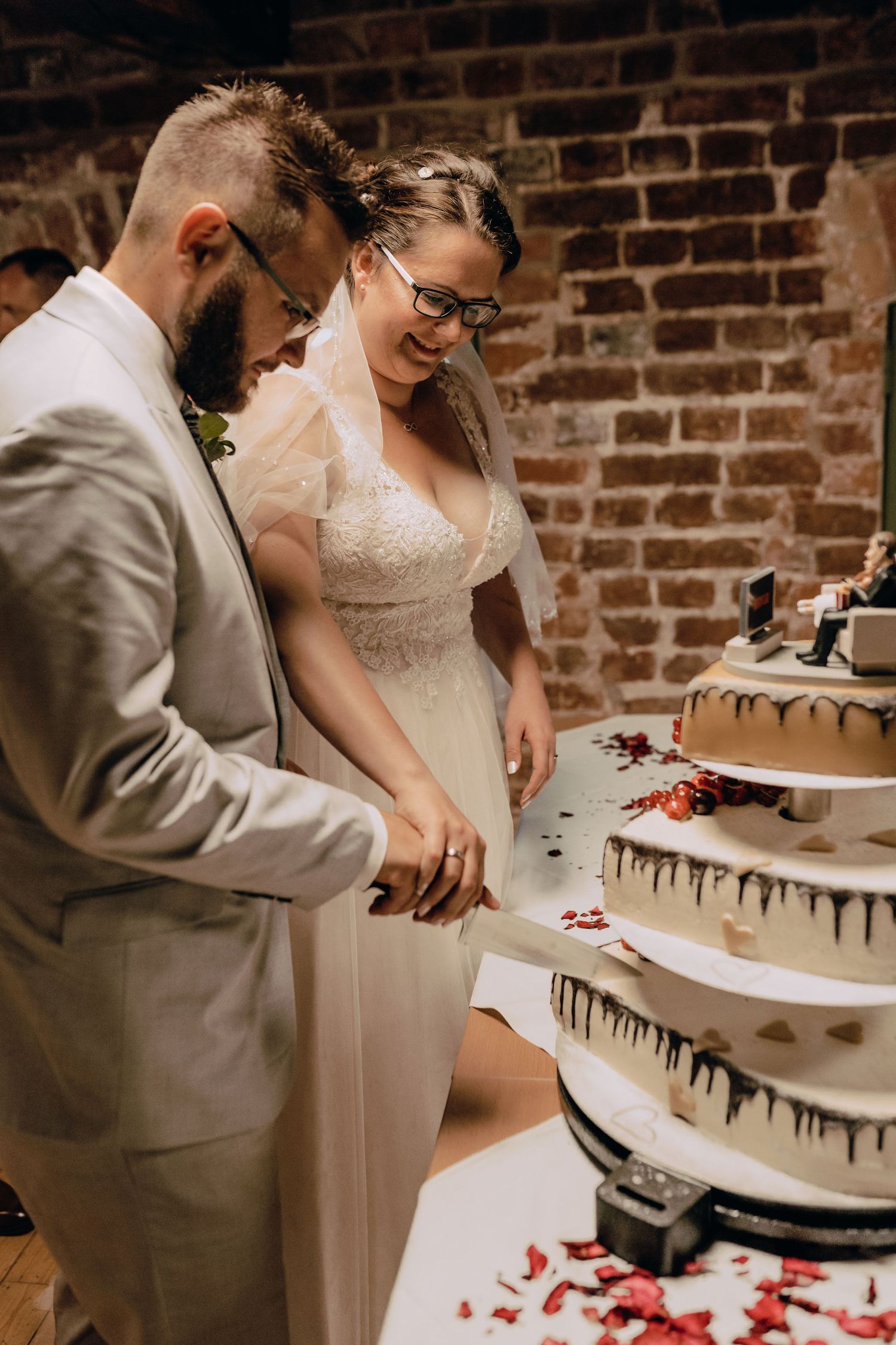 Couple cutting their beautiful black and white wedding cake decorated with red rose petals on a rustic table.