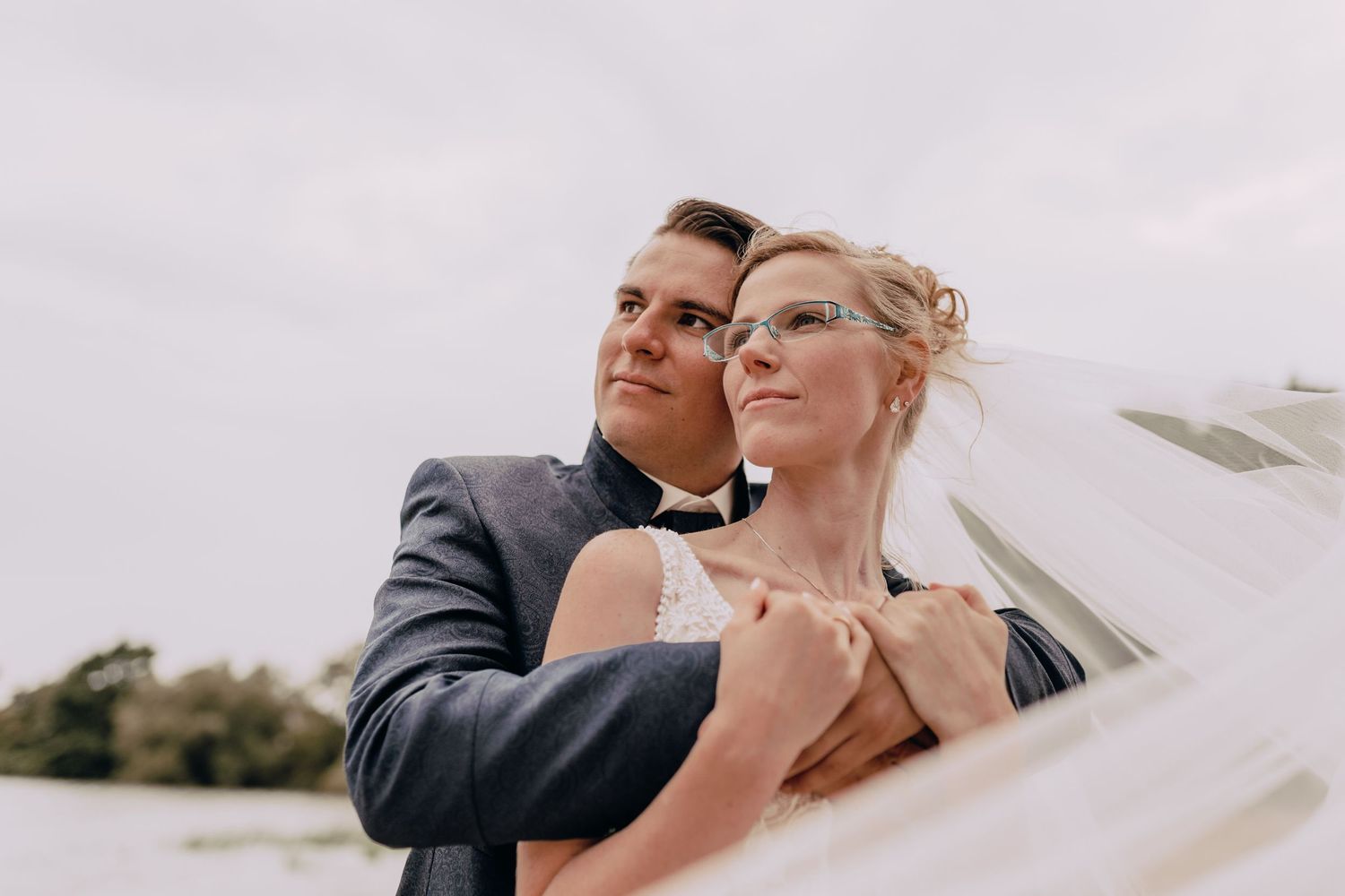 A bride and groom embrace outdoors on their wedding day with soft lighting and a dreamy veil flowing in the breeze.
