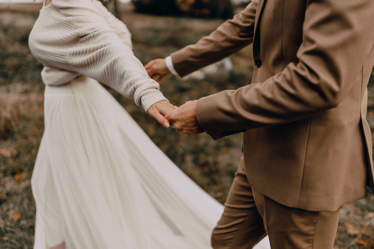 A romantic wedding couple in brown and cream outfits holds hands together in a nature setting.