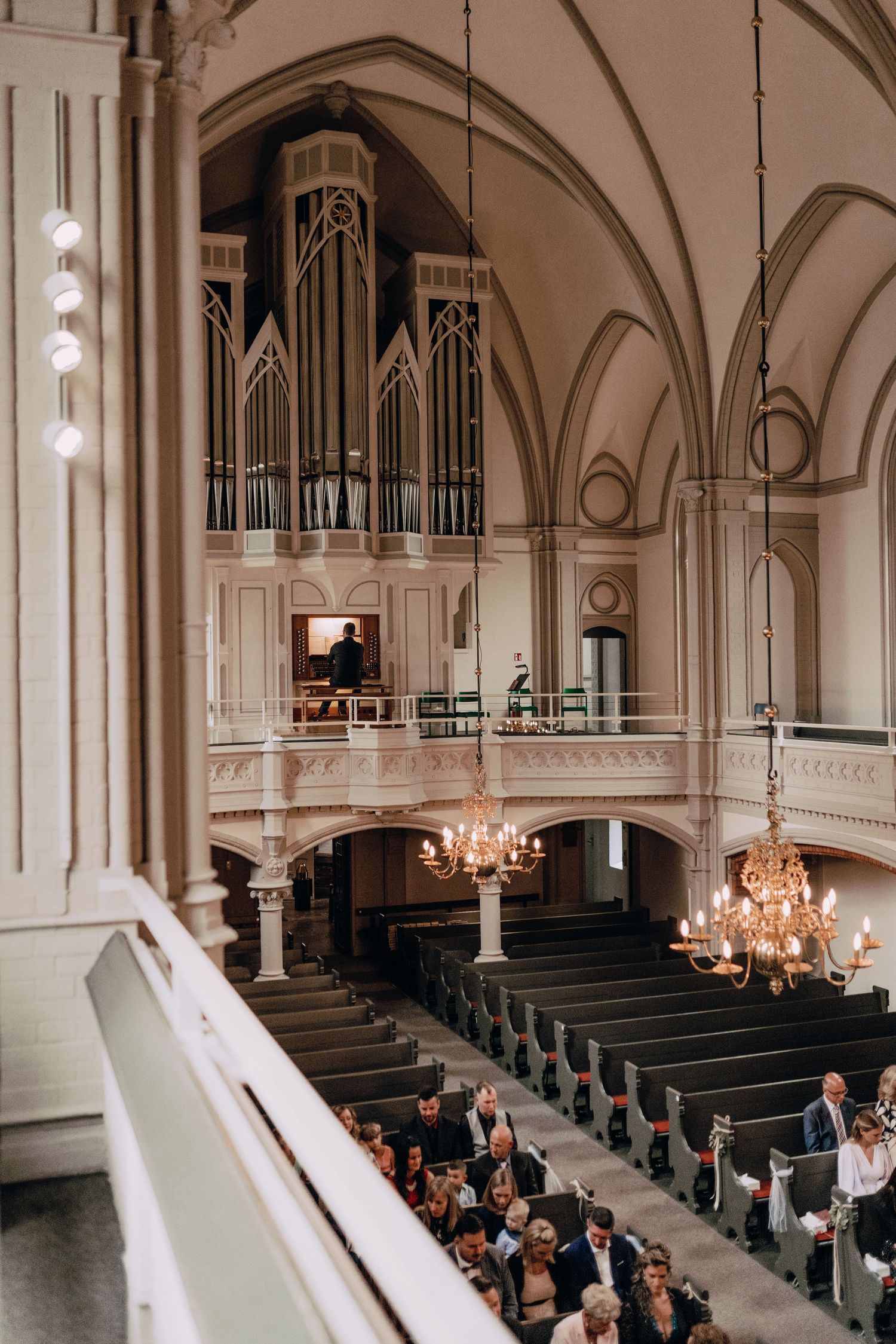 Interior of a historic church with a pipe organ, elegant chandeliers, and gathered attendees in traditional wooden pews.